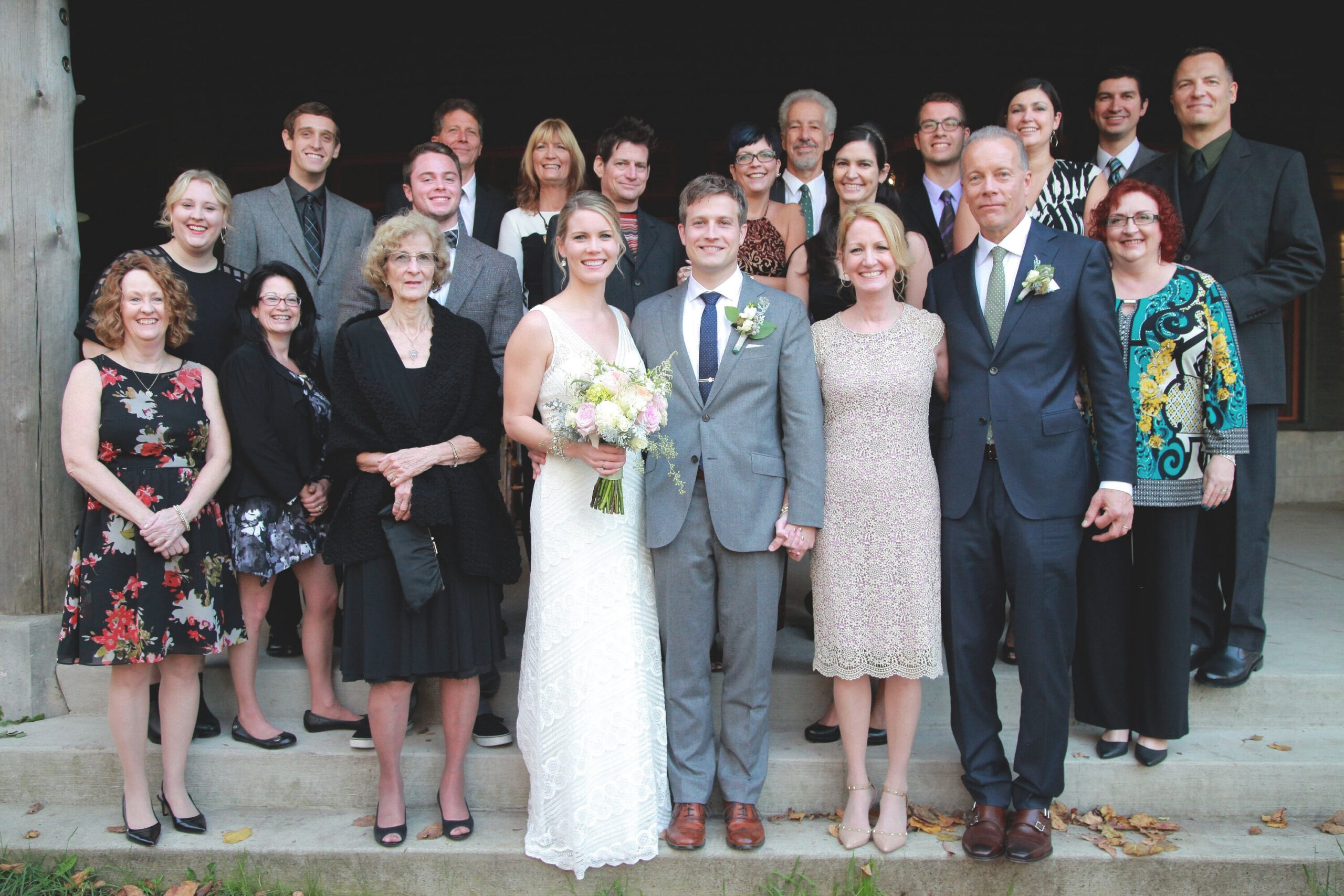 Wedding party posing on steps with bride in white gown and groom in gray suit at center, surrounded by family and friends.