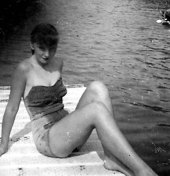 Woman in a strapless swimsuit sits on a dock by a lake, vintage black-and-white photo style.
