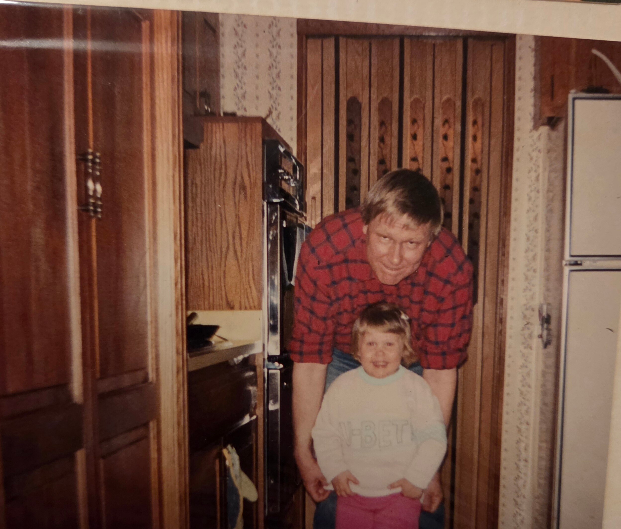 Family photo taken in a retro kitchen: a man in a red plaid shirt leaning over two children, with wood paneling and cabinets in the background.