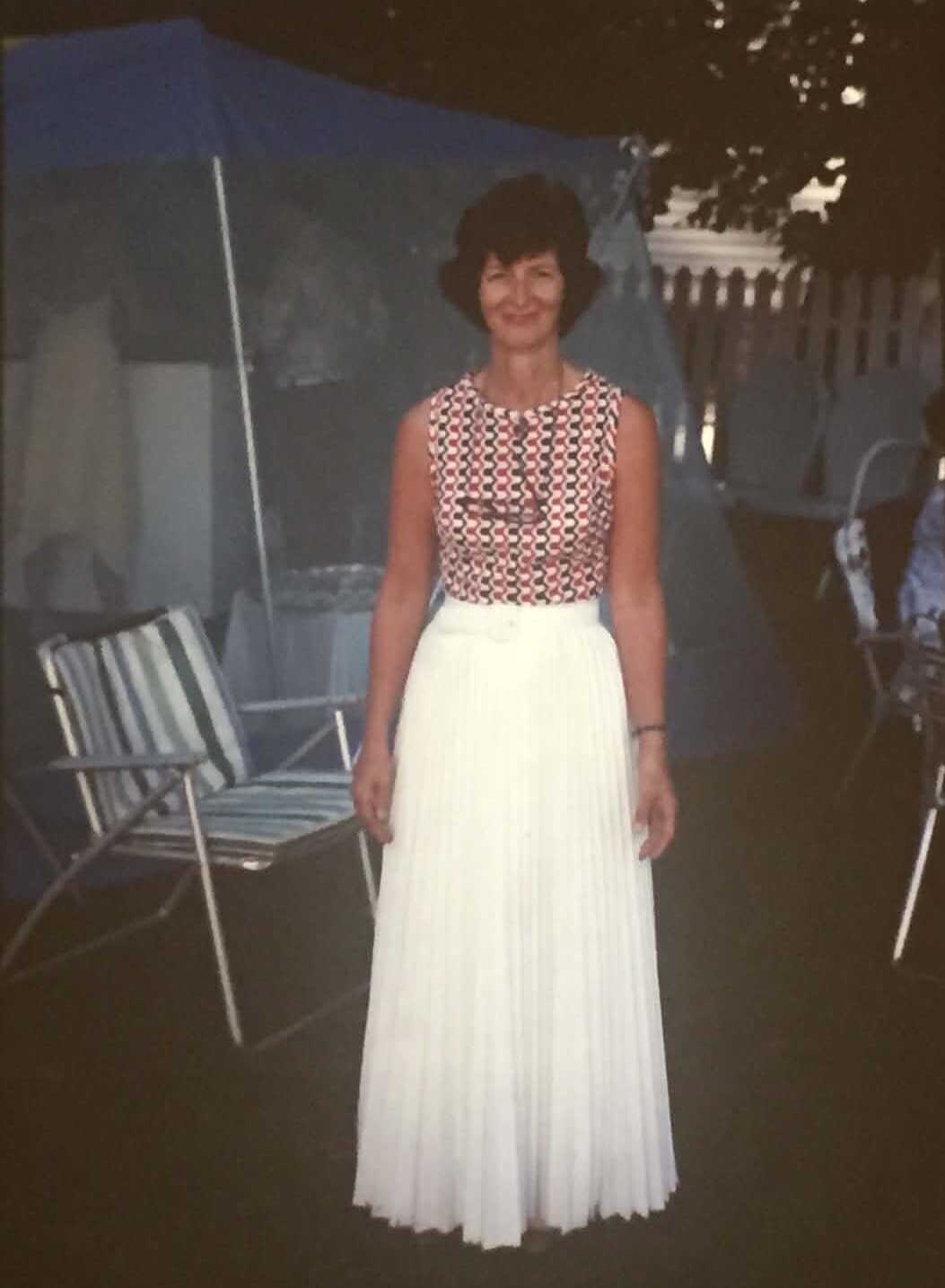 Woman posing outdoors in a sleeveless red‑white‑black patterned top and long white pleated skirt.