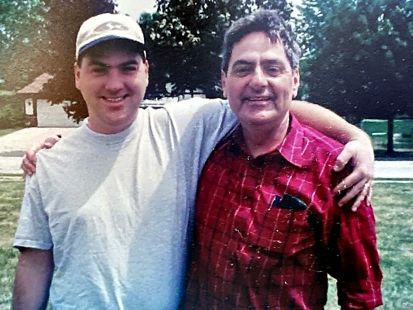 Two men stand side by side with arms around each other in a park, smiling at the camera.