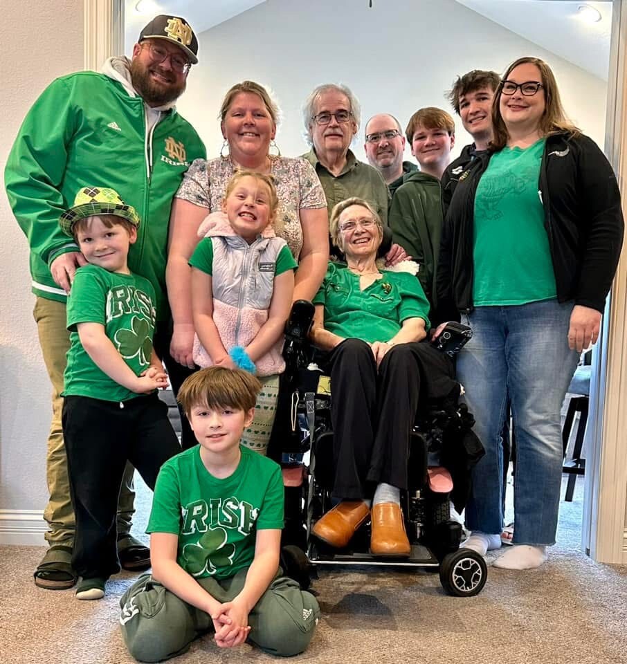 A smiling family group gathered in a doorway, centered around a woman in a motorized wheelchair, with children and adults wearing green Saint Patrick's Day attire.