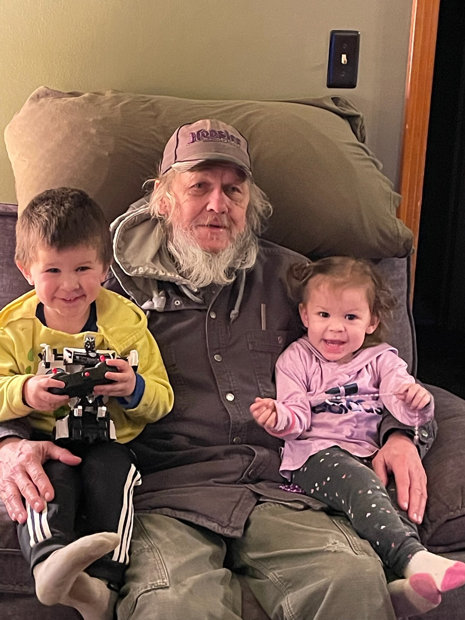 Older man with a white beard and cap sits on a couch between a smiling boy holding a toy vehicle and a girl in a pink shirt, all together on couch cushions.