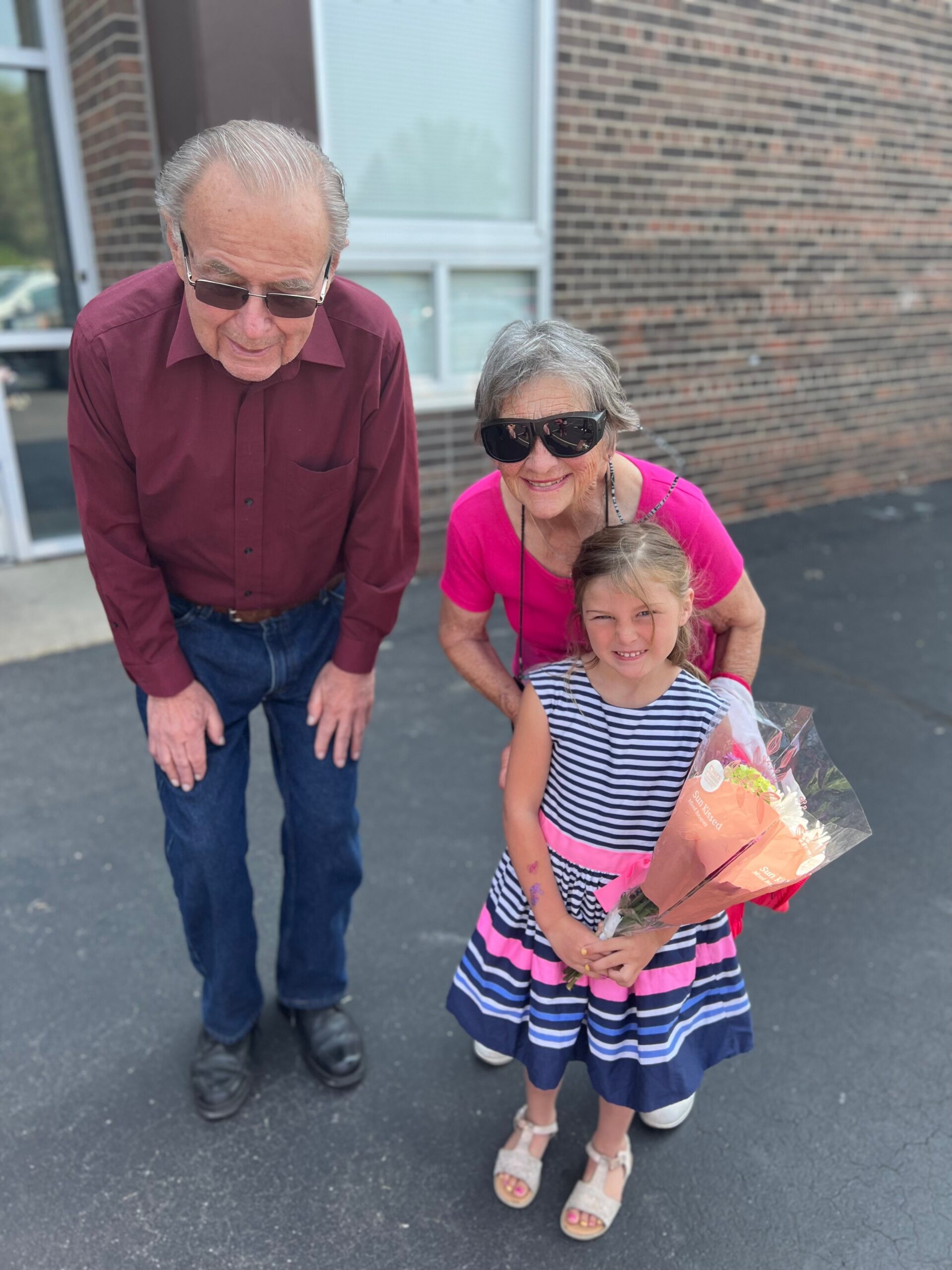 Grandfather, grandmother, and granddaughter pose together outside a brick building; the girl holds a bouquet of flowers.