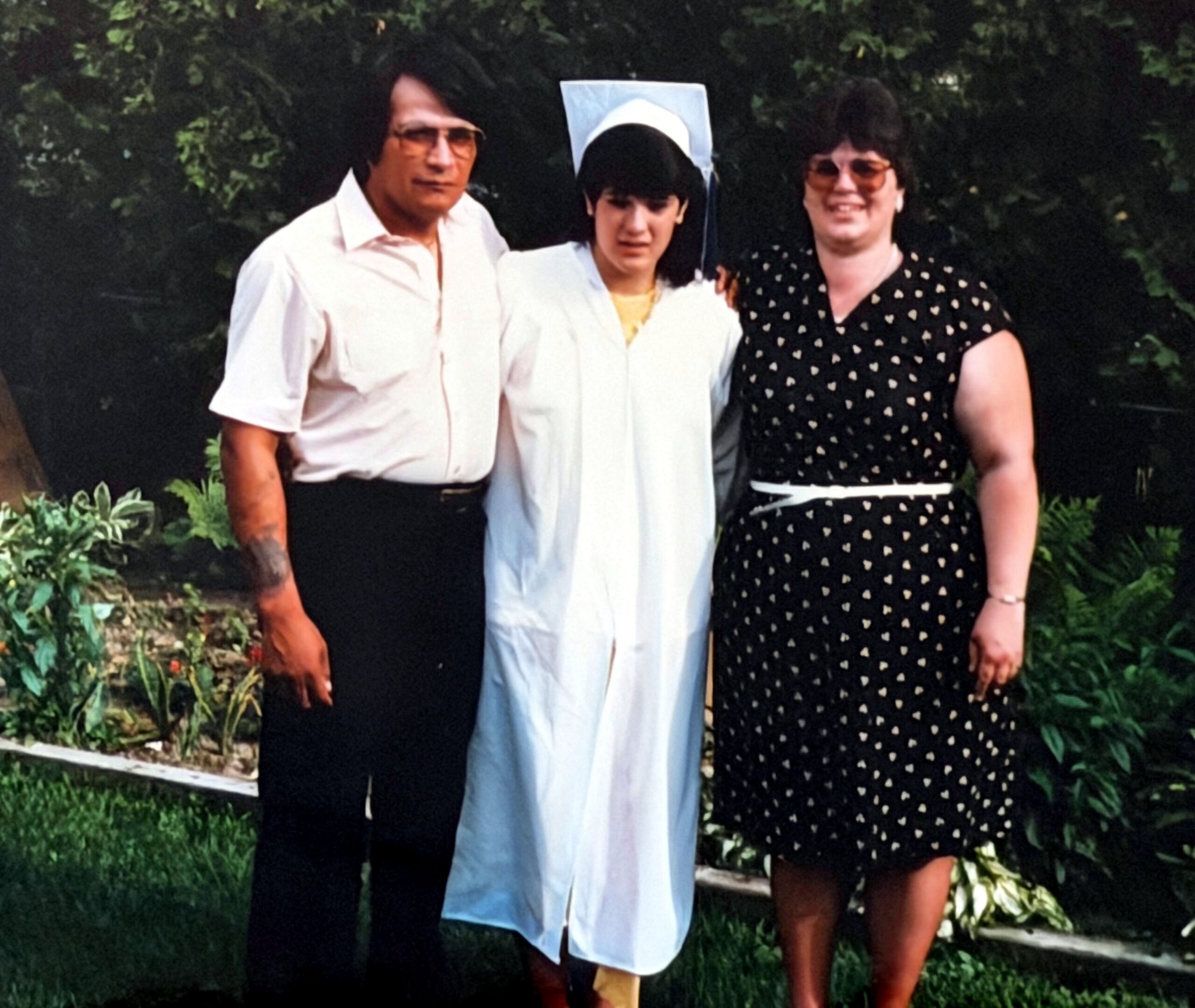 Group photo of three people outdoors; a graduate in a light blue gown and cap stands between an older man in a white shirt and a woman in a black dress with small heart patterns in a garden setting.