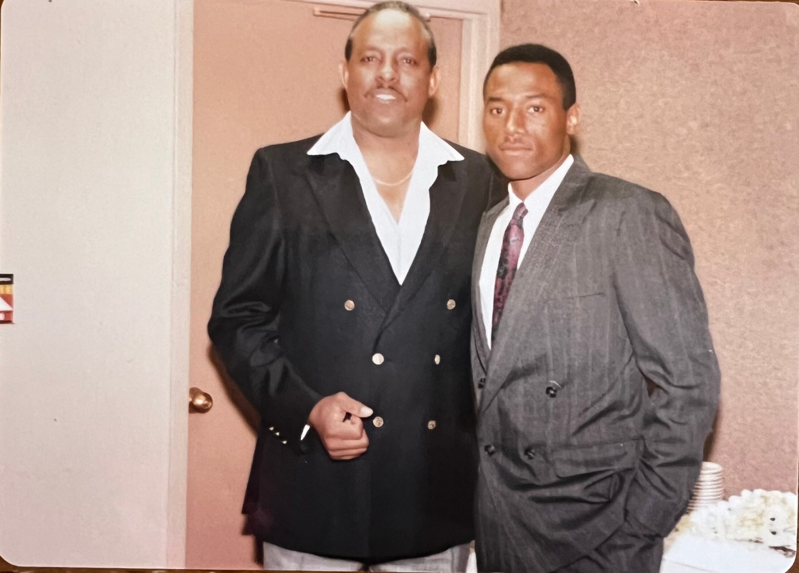 Two men in suits posing indoors, one in a black double-breasted blazer and the other in a gray pinstripe suit.