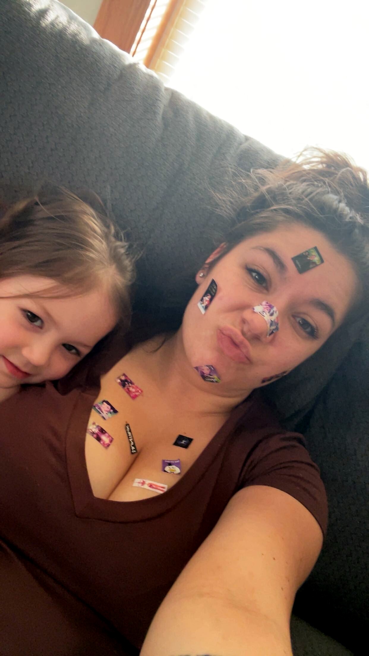 Woman taking a playful selfie on a dark couch; stickers stuck to her face and chest while a young child sits beside her, smiling slightly beside them.