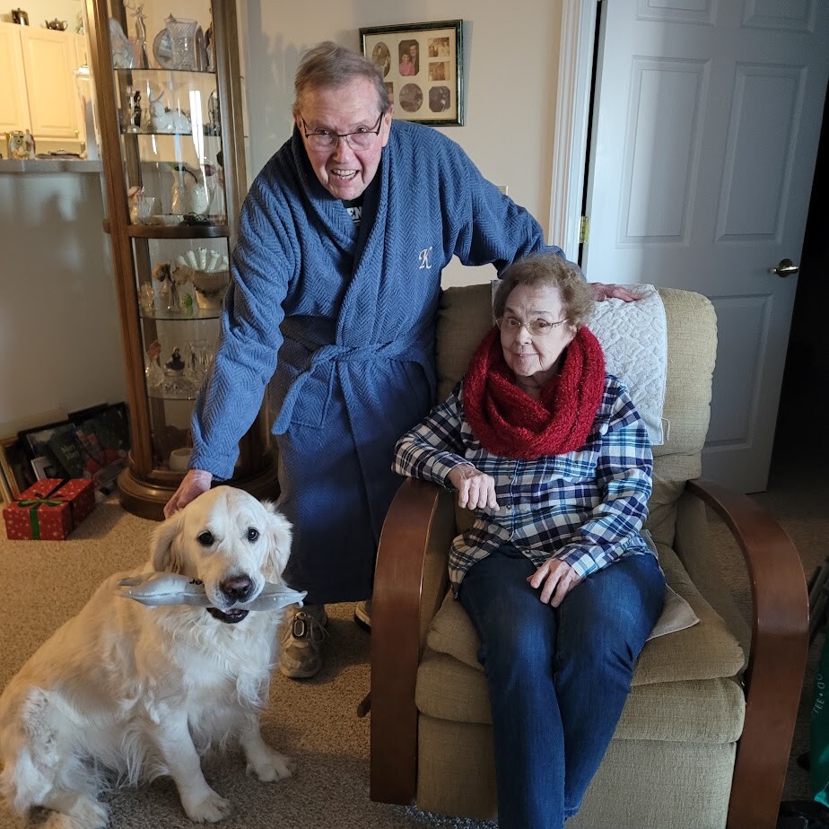 An elderly woman in a plaid shirt and red scarf sits in a beige chair while a man in a blue robe leans over smiling; a golden retriever sits on the floor with a toy in its mouth nearby.