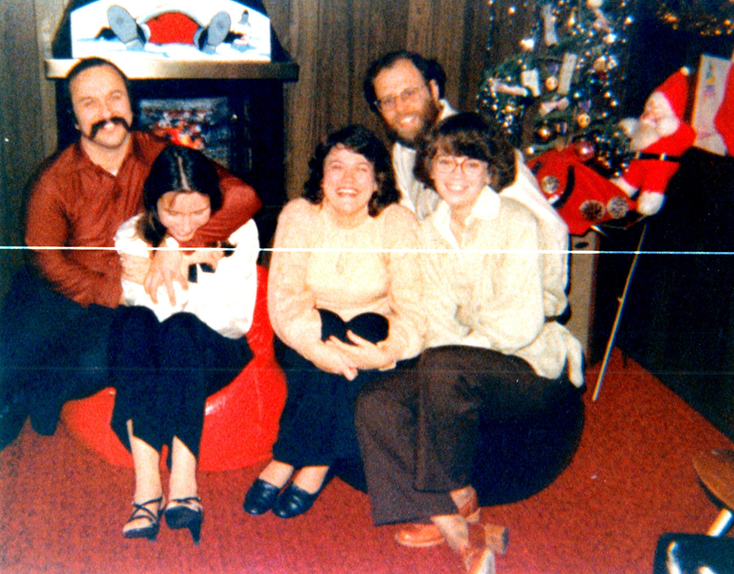 Group of five adults posing for a holiday photo in a living room with a Christmas tree and a Santa figure in the background.