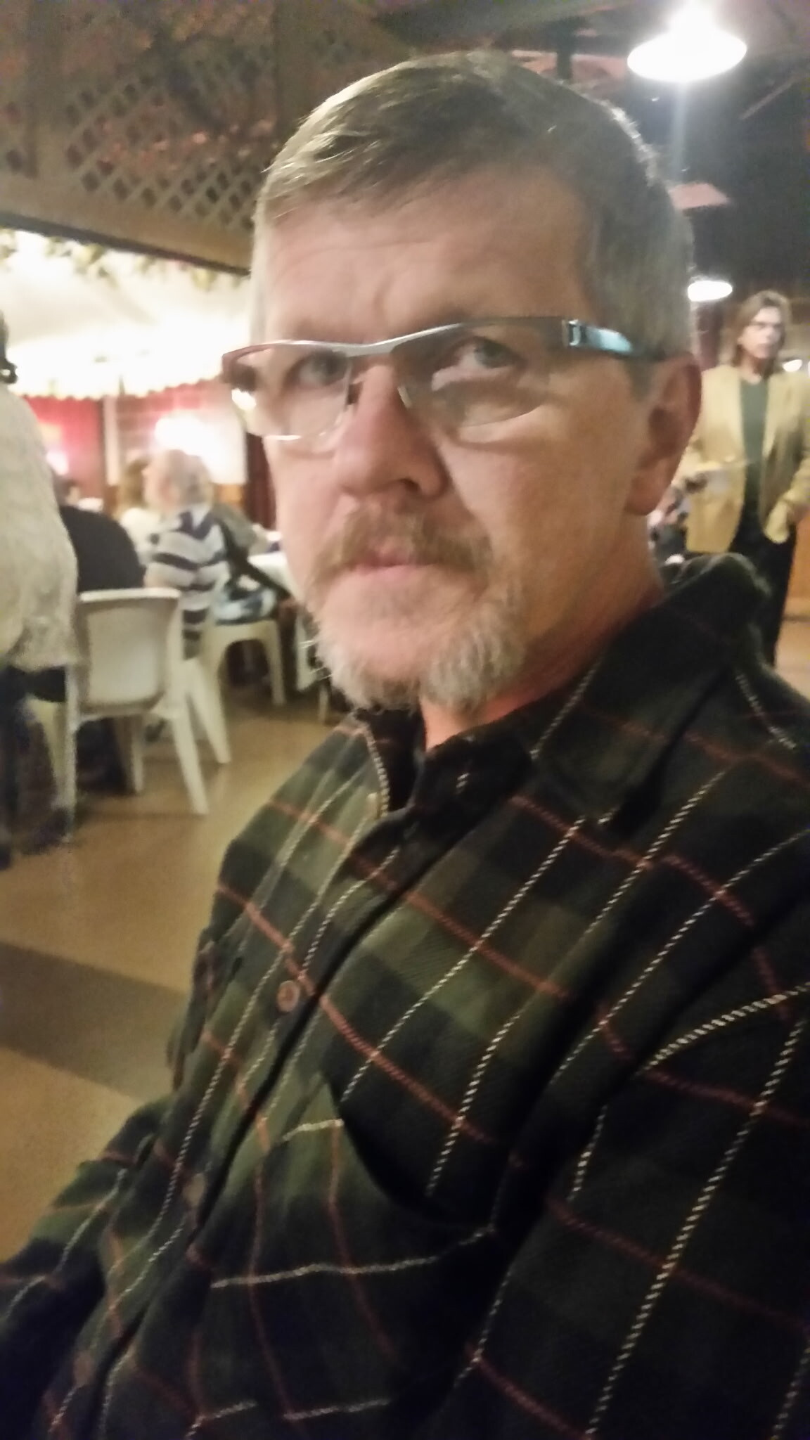 Close-up of a man with short gray hair and beard wearing glasses and a green plaid shirt, seated in a restaurant with other diners in the background.