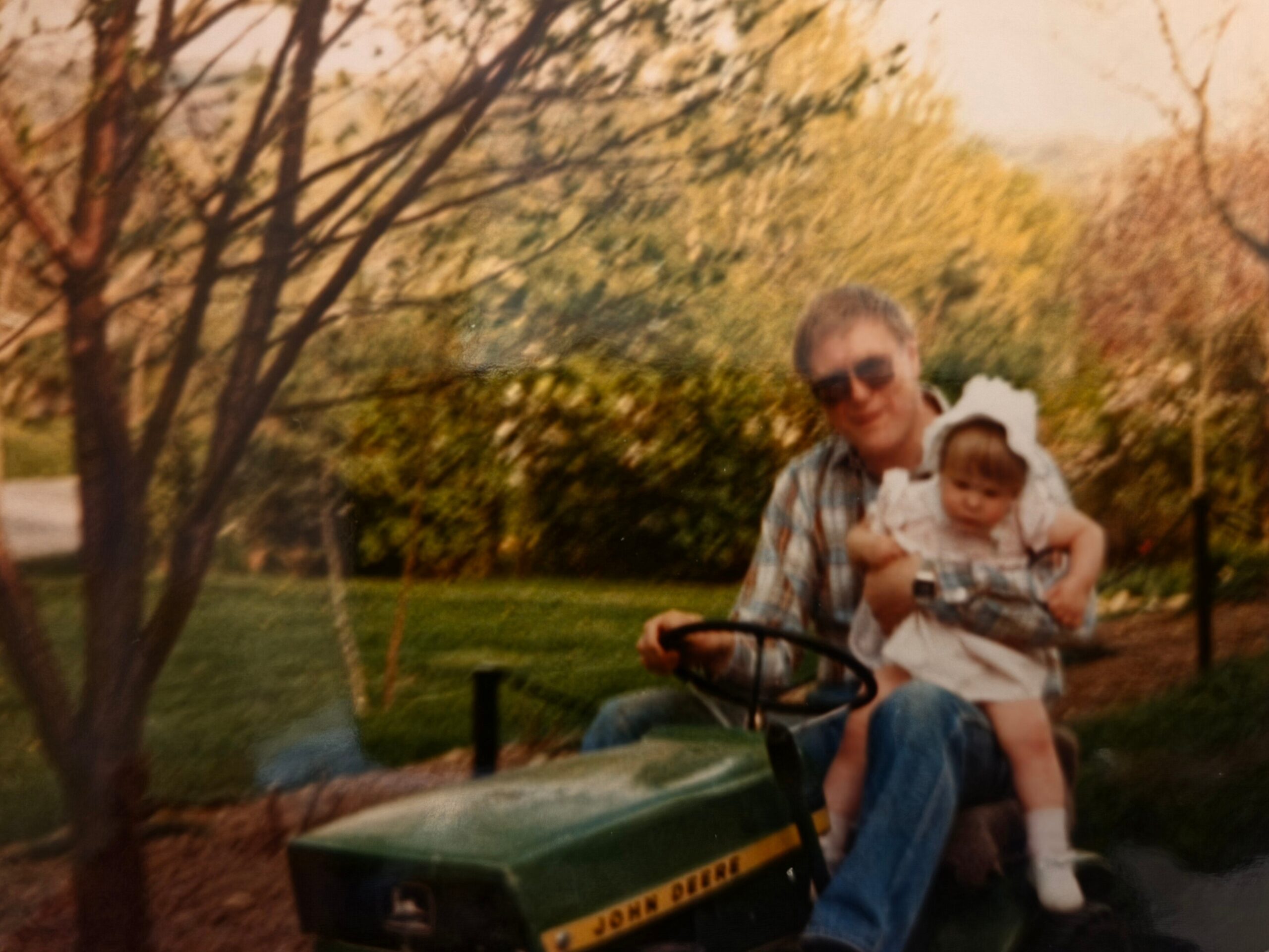 Man wearing sunglasses rides a green John Deere pedal tractor with a baby in his arms, outdoors among trees.