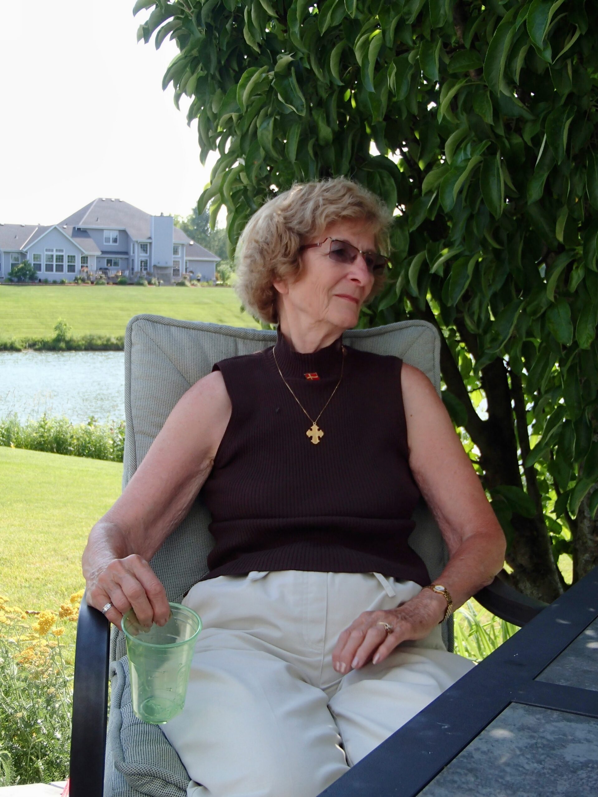 Older woman relaxing outdoors in a garden, sitting in a chair with a green plastic cup in her hand.