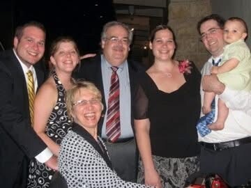 Smiling family group posing for a photo at a celebration, with seven adults and a baby.