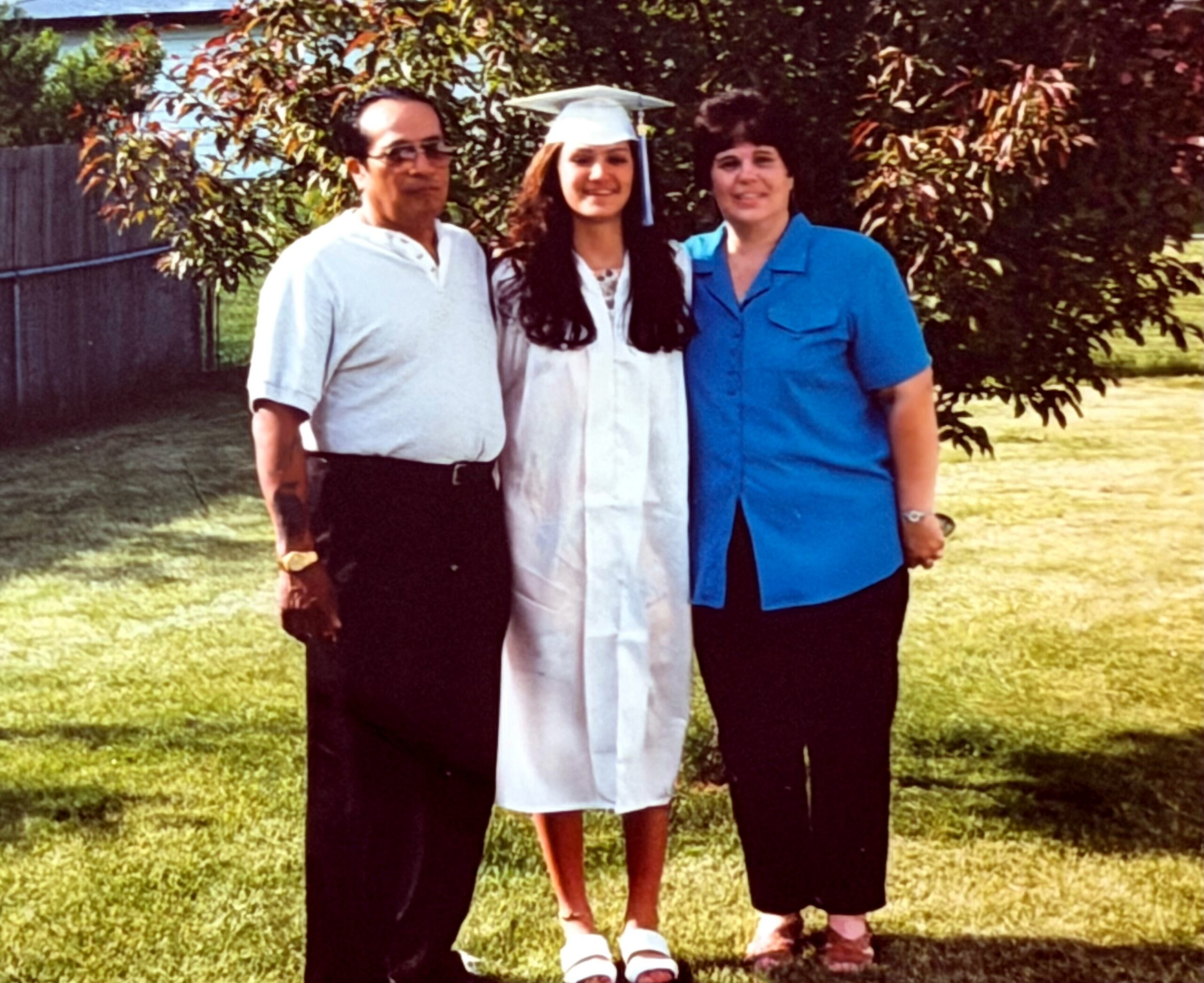Graduate in a white gown and cap stands between a man in a light shirt and a woman in a blue shirt outdoors in a yard with trees in the background.