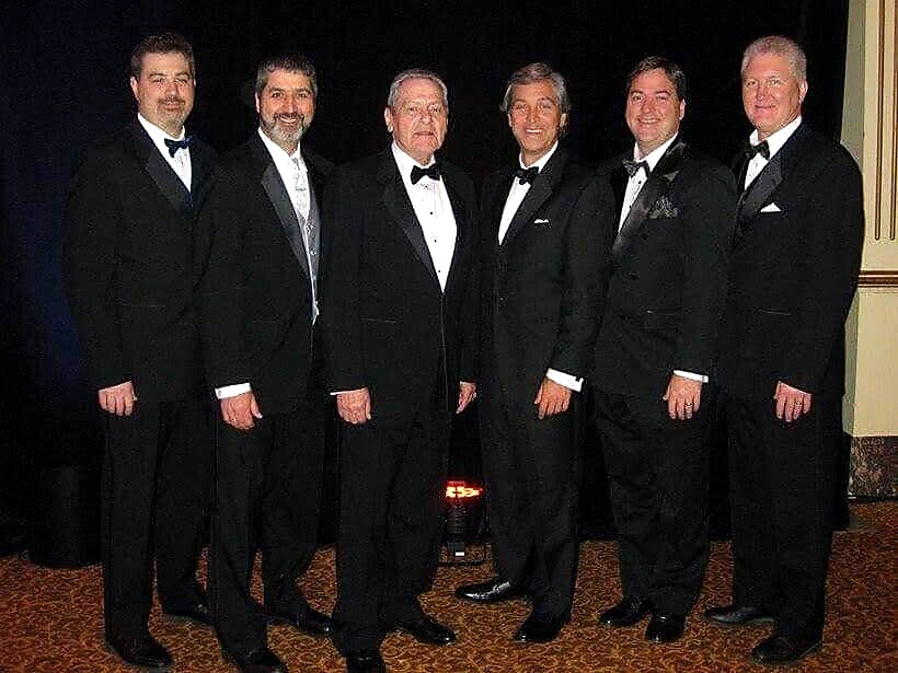 Seven men in black tuxedos posing together for a group photo at an event, smiling at the camera.