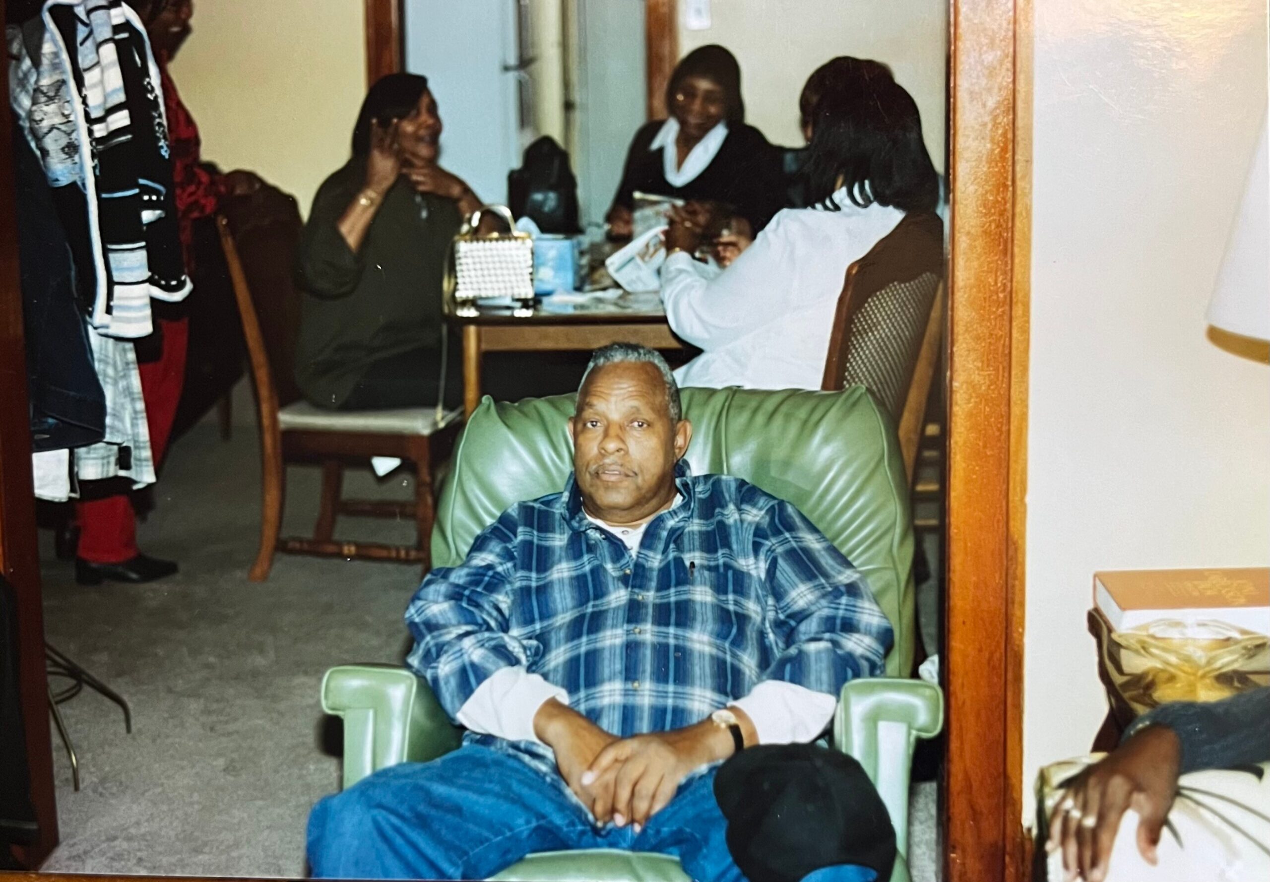 Man in a blue plaid shirt relaxing in a green armchair, with friends sitting and chatting at a table in a home setting behind him.