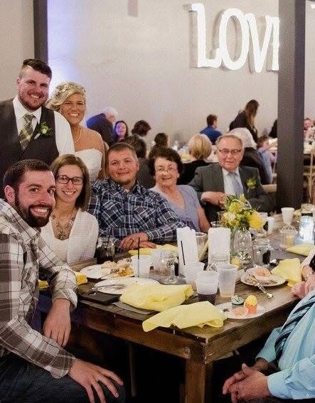 Group of wedding guests around a table, smiling at the camera, with a bride and groom standing behind them in a banquet hall with a 'LOVE' sign in the background.