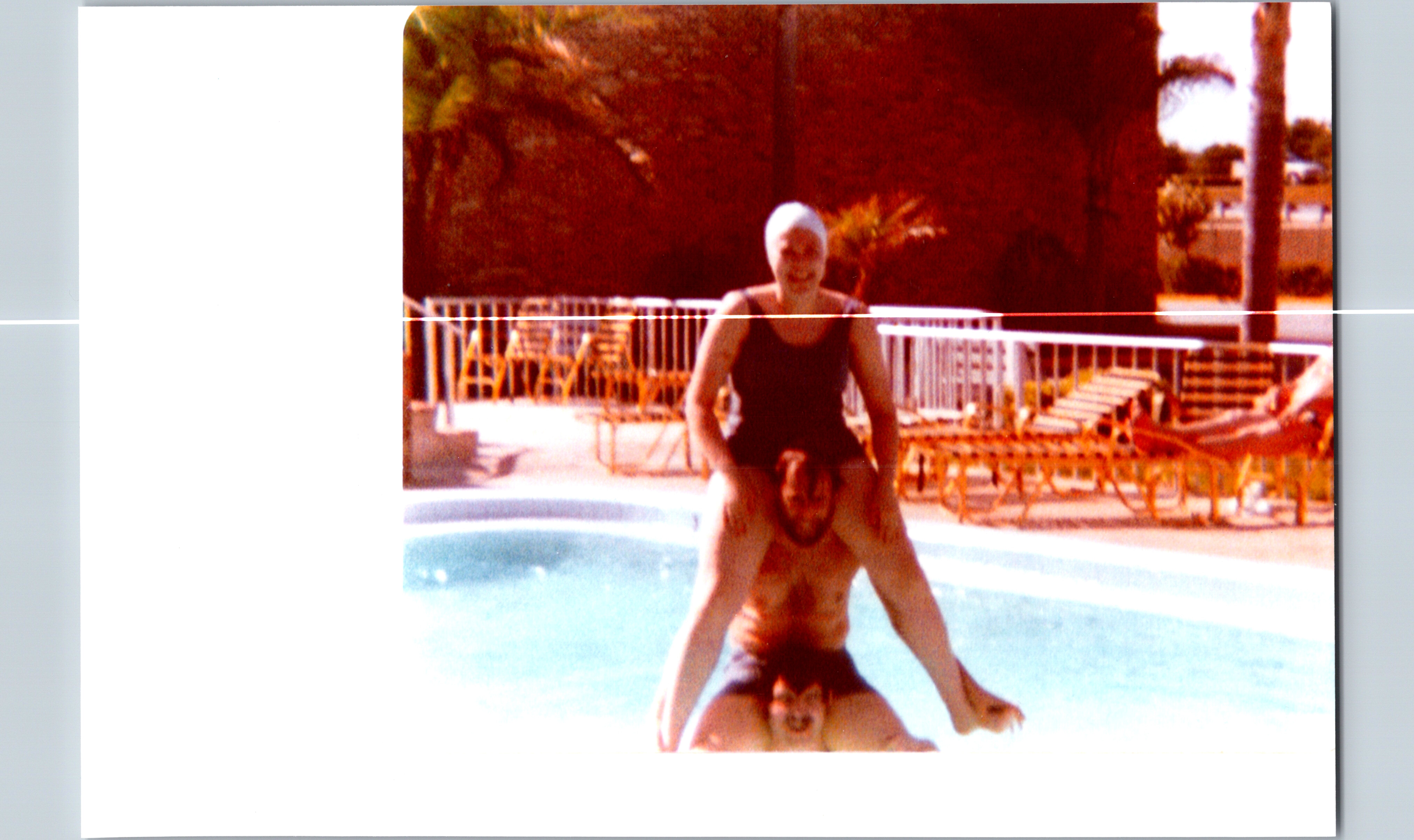 Person wearing a white swim cap sitting on another person’s shoulders at a poolside scene with loungers in the background.