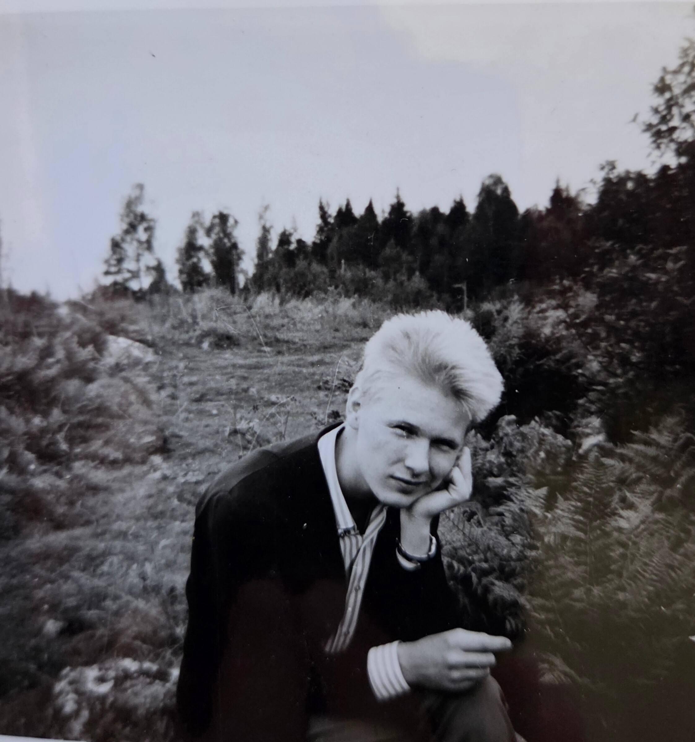 Black-and-white photo of a man outdoors in a field, with trees in the background and a fluffy white dog partially in the foreground.