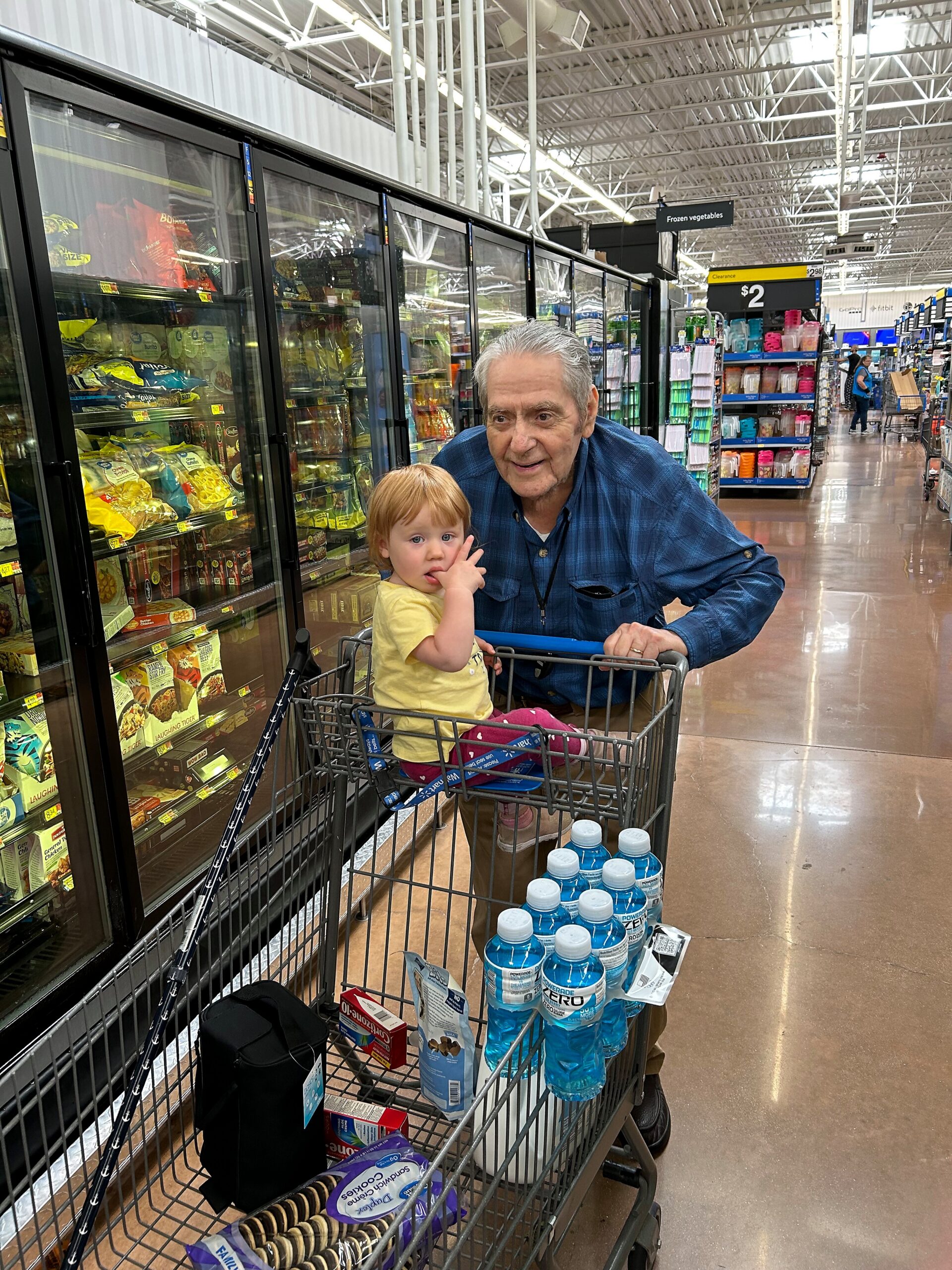 An elderly man leans over a shopping cart with a young child inside, near frozen food freezers in a supermarket aisle.