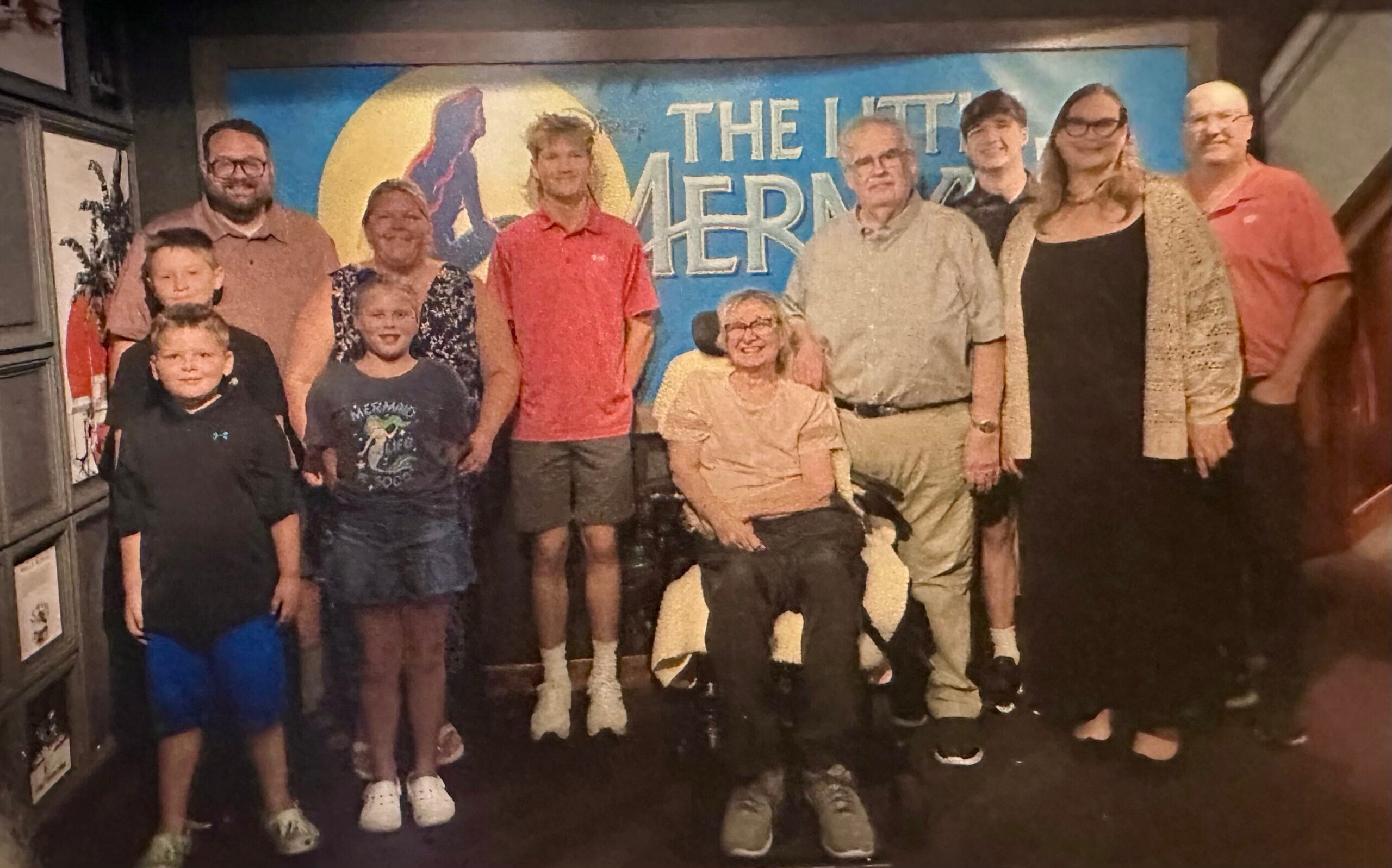 Group of ten people posing for a photo in a theater lobby, with a blue mural behind them that reads 'THE LITTLE MERMA...' and a central woman in a wheelchair among adults and children smiling at the camera.