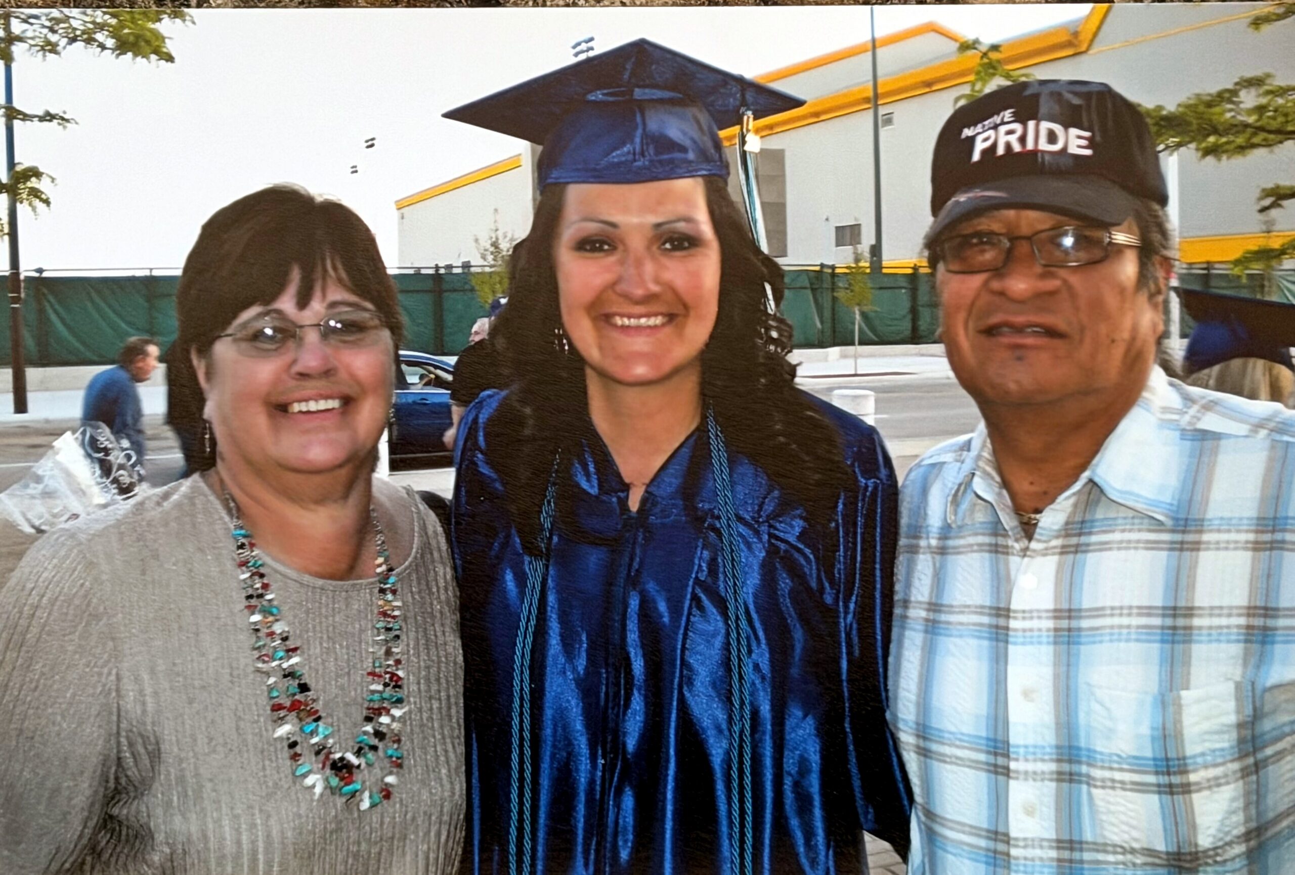 Three smiling adults posing outdoors; the center person wears a blue graduation gown and cap, celebrating graduation with two companions beside her.