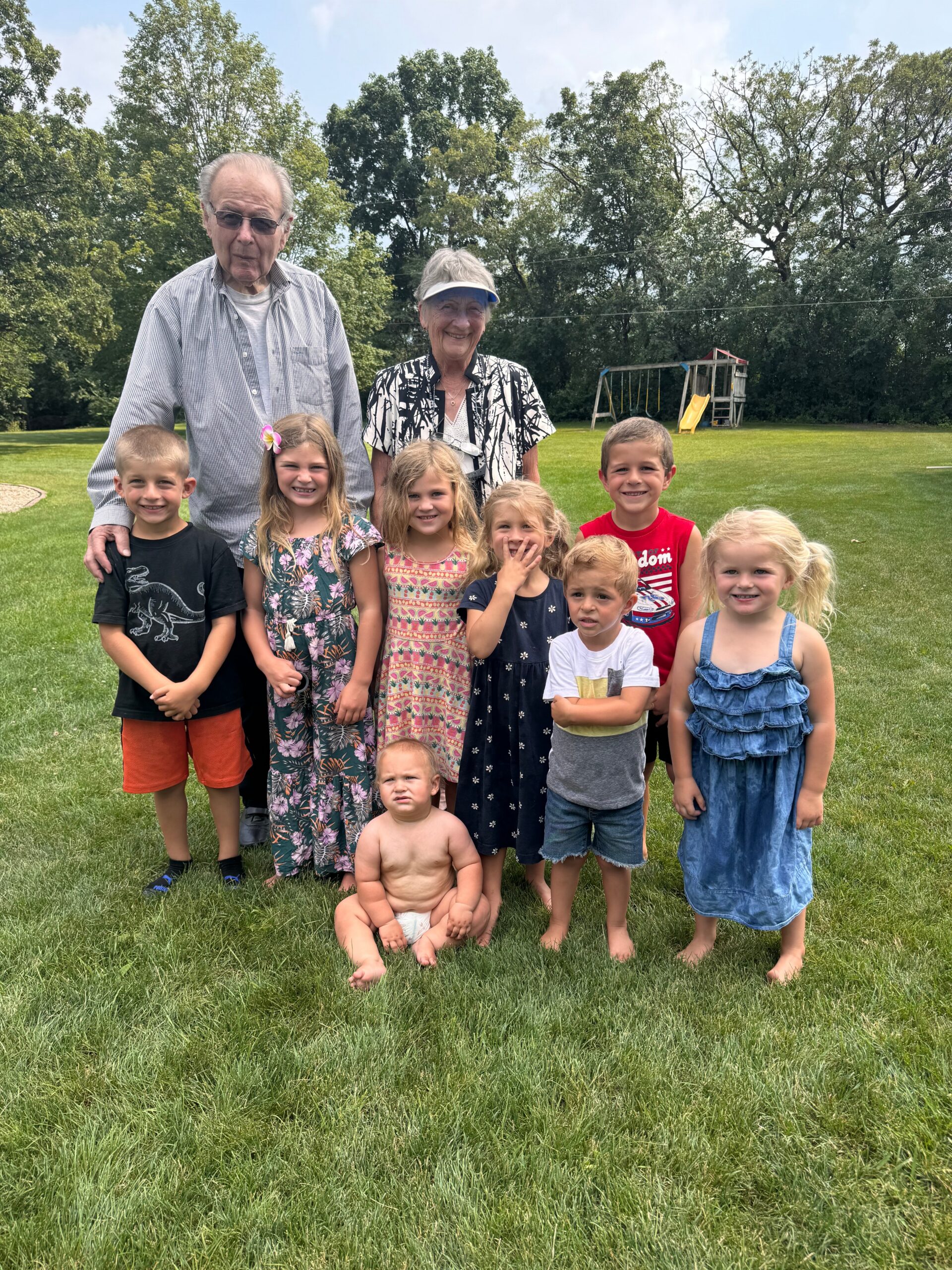 Group of children and two adults posing on a grassy park lawn with trees and a playground in the background.