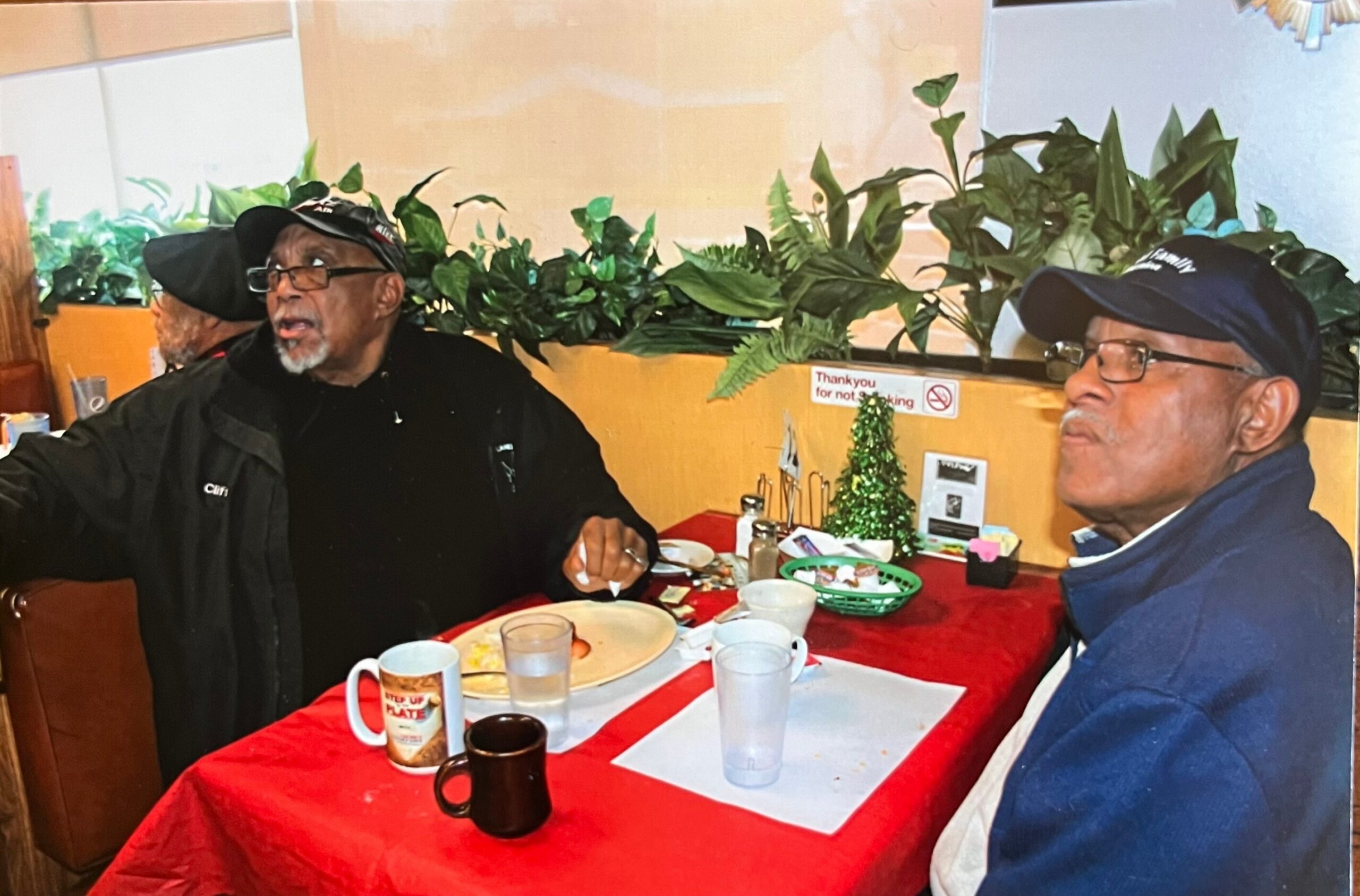 Two men sit at a red-clothed table in a cafe, with dishes and mugs, and a green plant backdrop behind them.