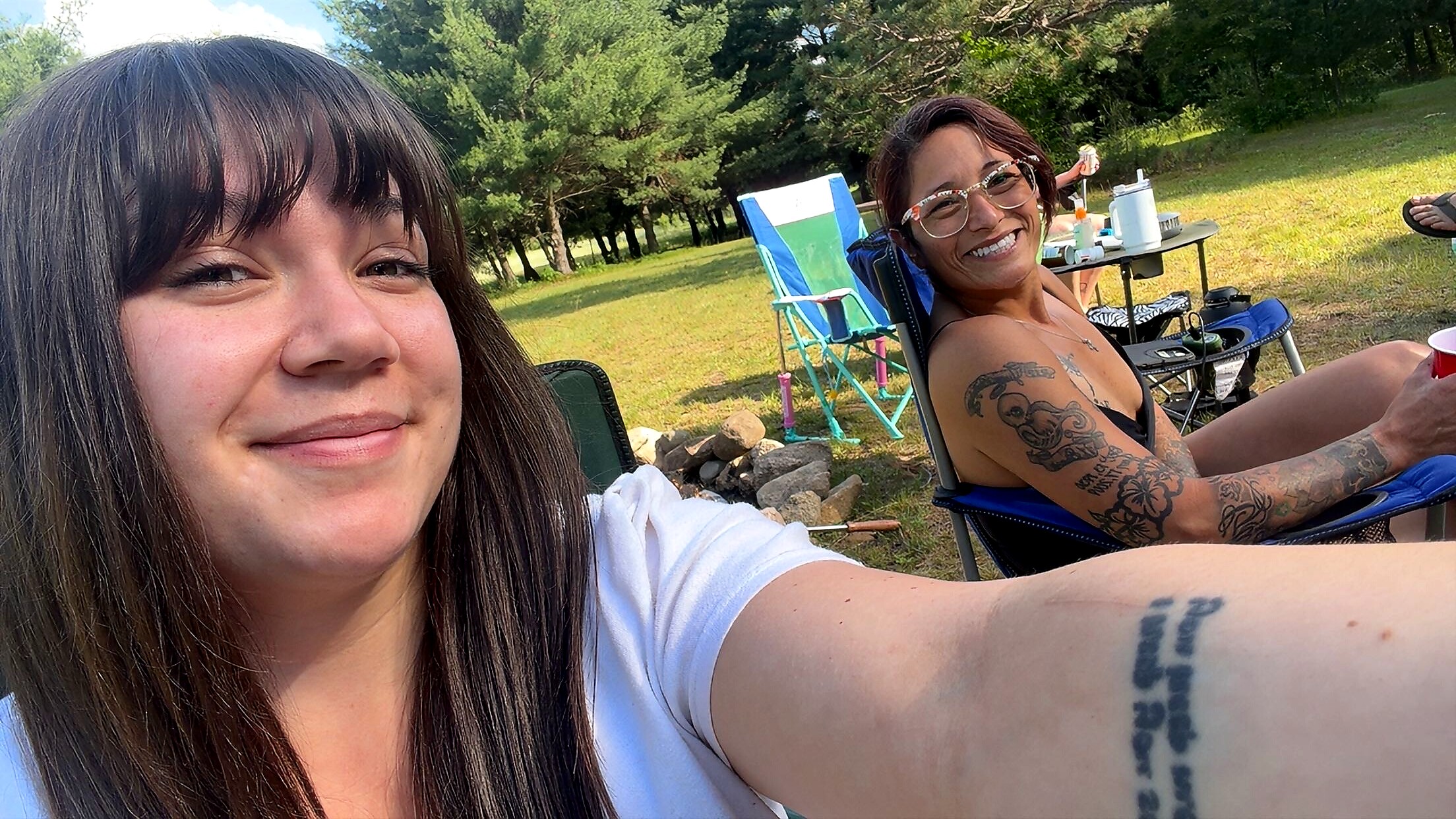 Two smiling women relaxing at a sunny outdoor picnic, one taking a selfie with tattoos on her arm visible.