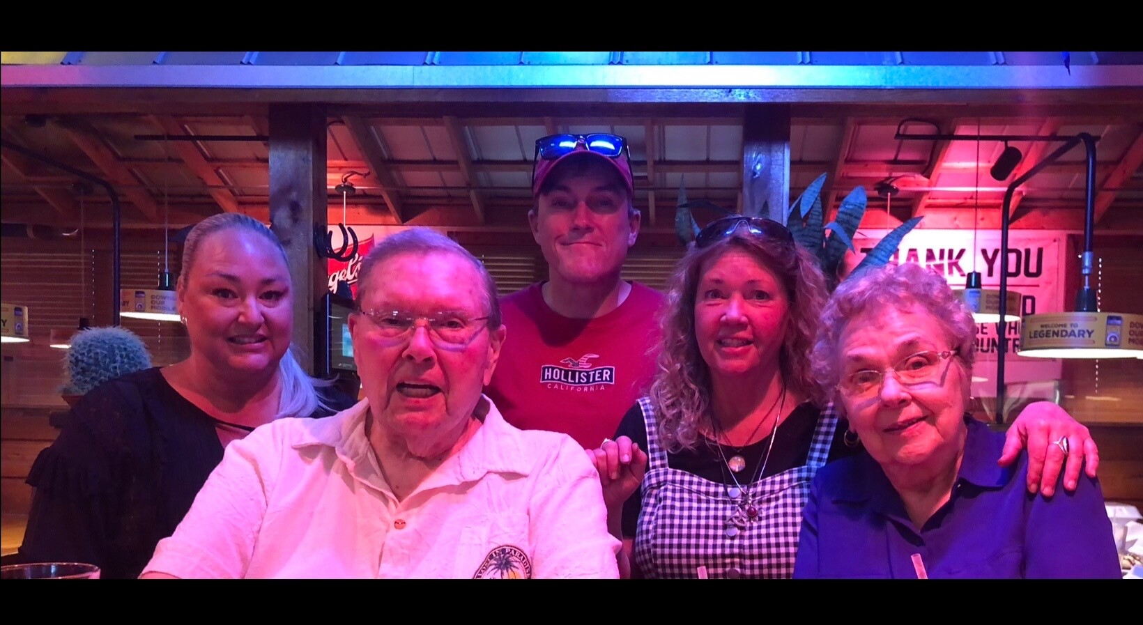 Five friends or family members pose together and smile at a casual restaurant or bar with colorful neon lighting and a wooden interior.