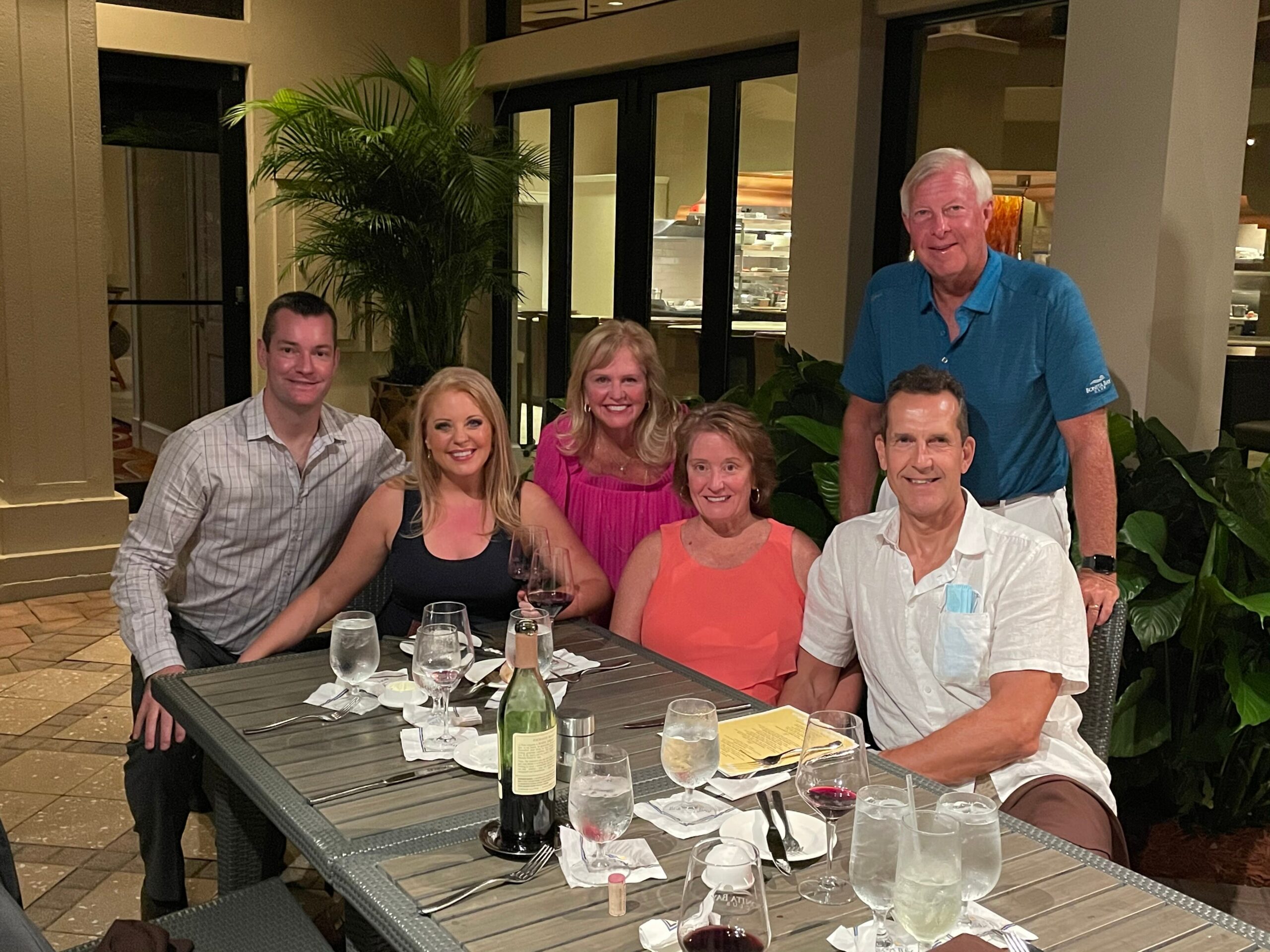 Six adults posing for a group photo at a restaurant table with wine glasses and a bottle visible, smiling at the camera.