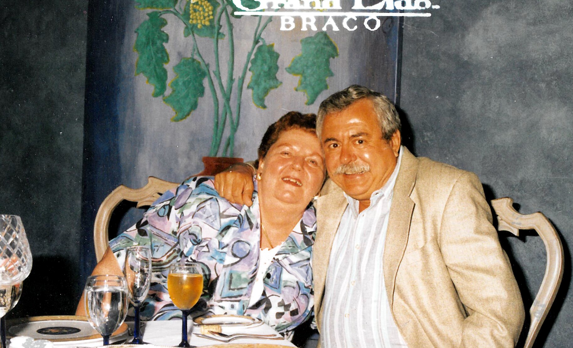 An elderly couple seated at a dining table, smiling for the camera with drinks in front of them and a plant mural behind them.
