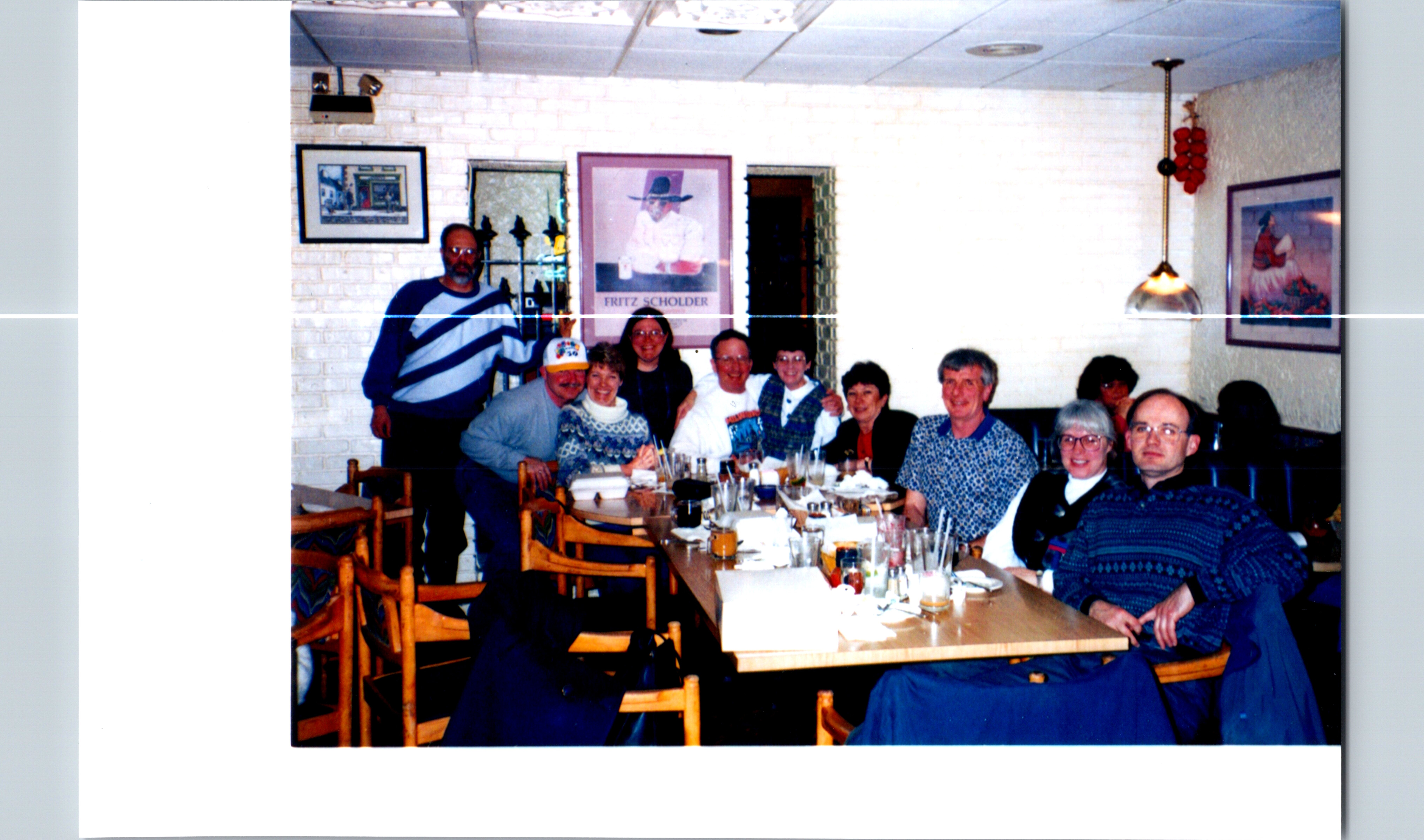 Group of adults gathered around a long dining table in a casual restaurant, smiling for the photo with plates and drinks on the table.