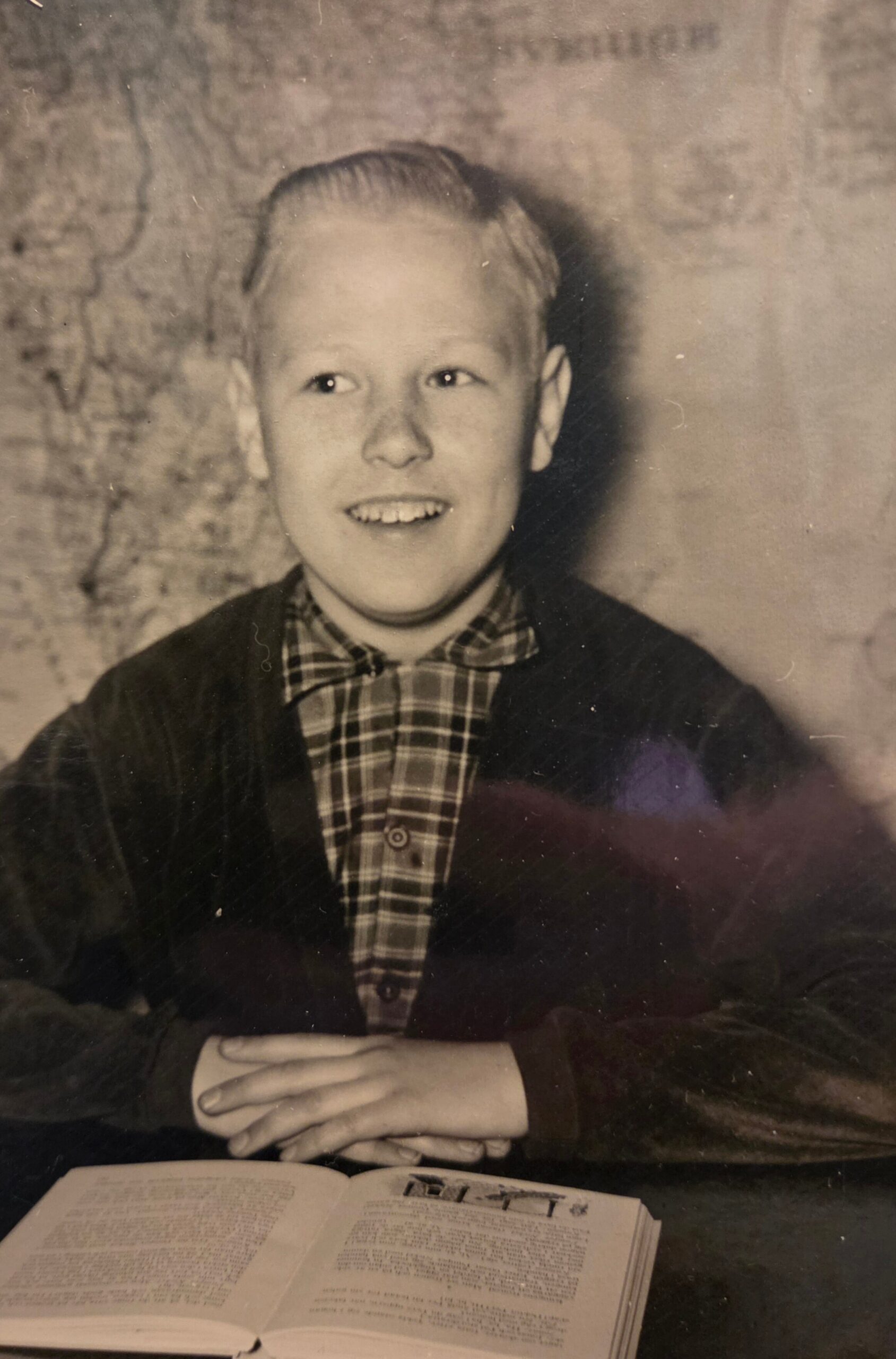 Smiling young boy in plaid shirt and dark cardigan sits at a table with hands folded, an open book in front of him (vintage photo).