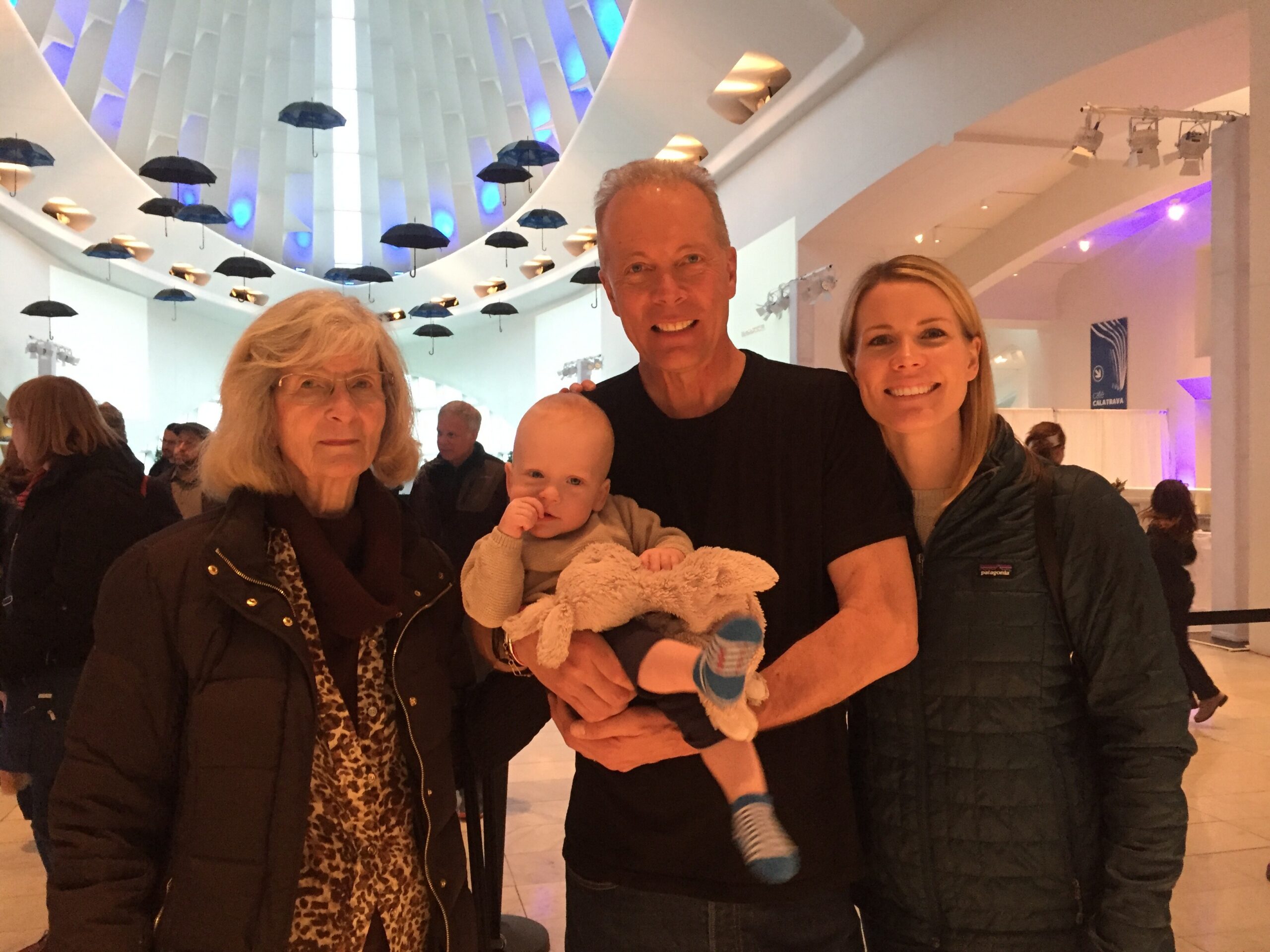 Group of four people posing indoors under a ceiling with hanging black umbrellas; man holds baby, two women beside him, all smiling.