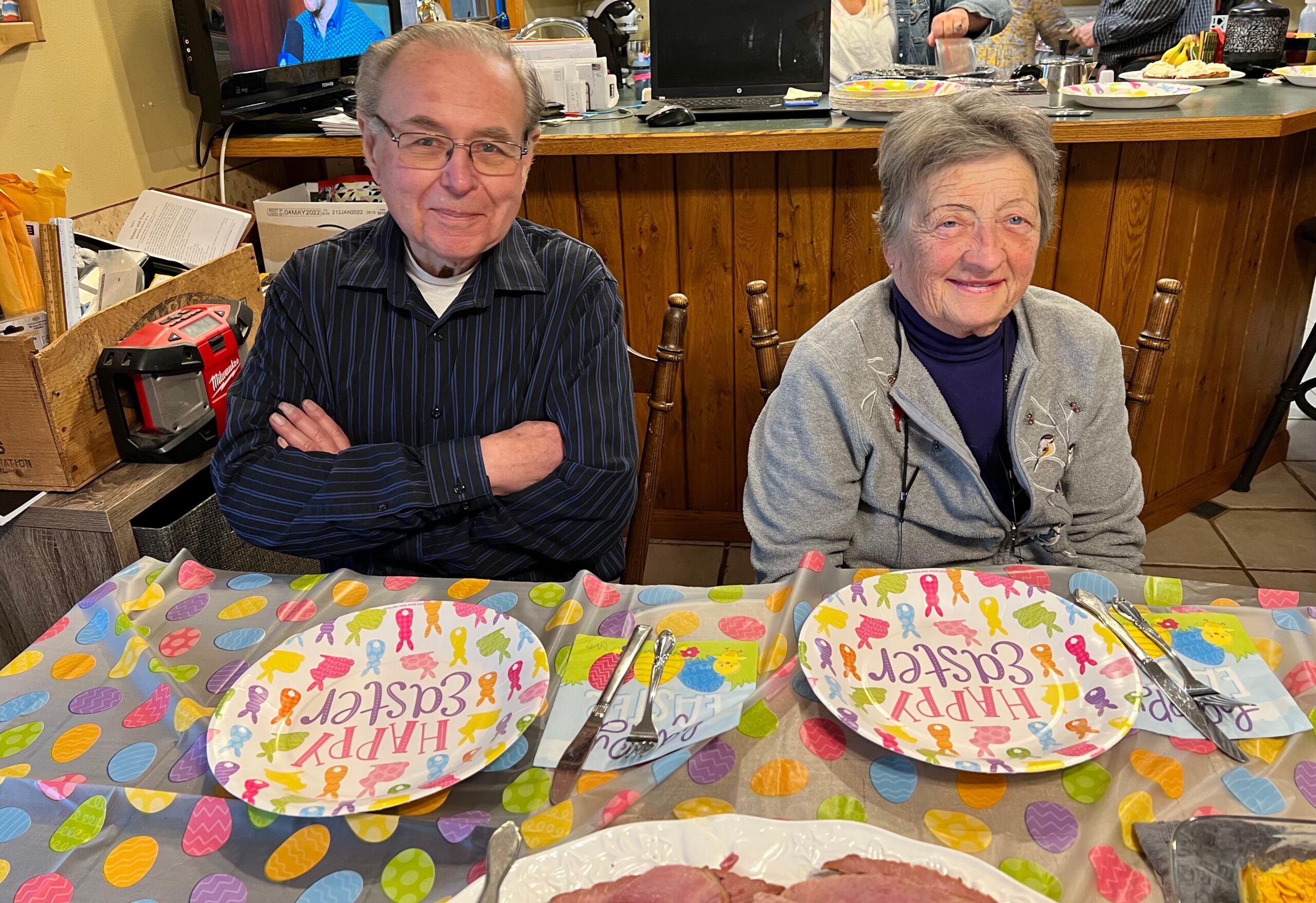 Two senior adults sit at a party table with colorful, patterned plates, smiling at the camera indoors.