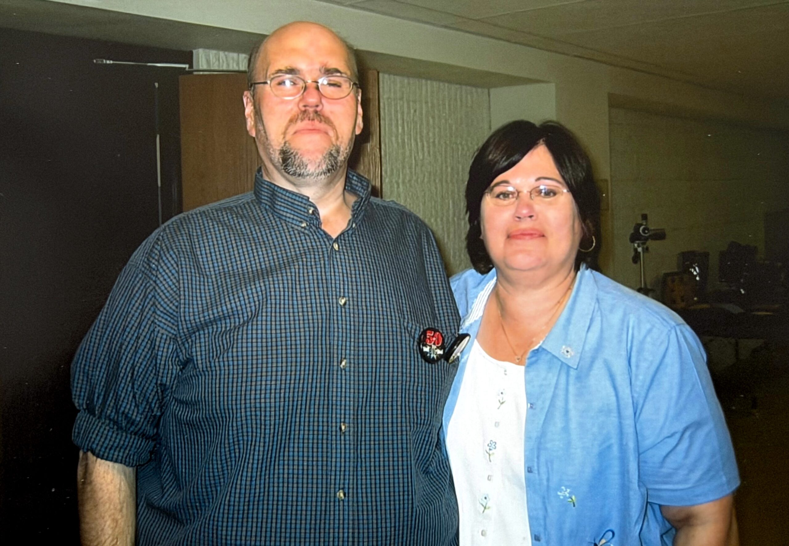 Two adults posing together indoors: a balding man with glasses and a goatee in a blue checkered shirt, and a woman with short dark hair and glasses wearing a light blue jacket nearby.