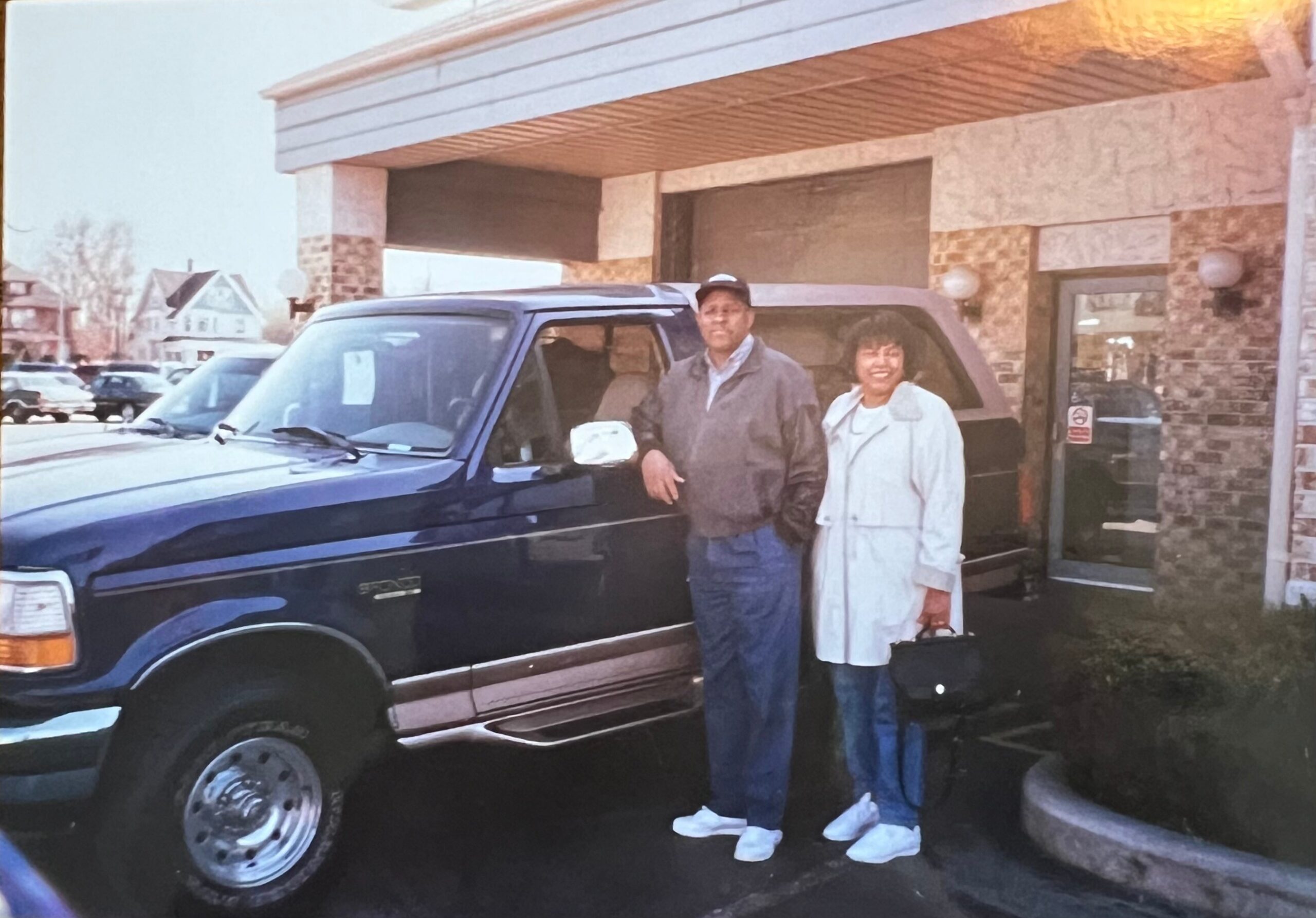 Two people stand beside a dark SUV in front of a storefront, posing for the photo outside a building with stone walls.