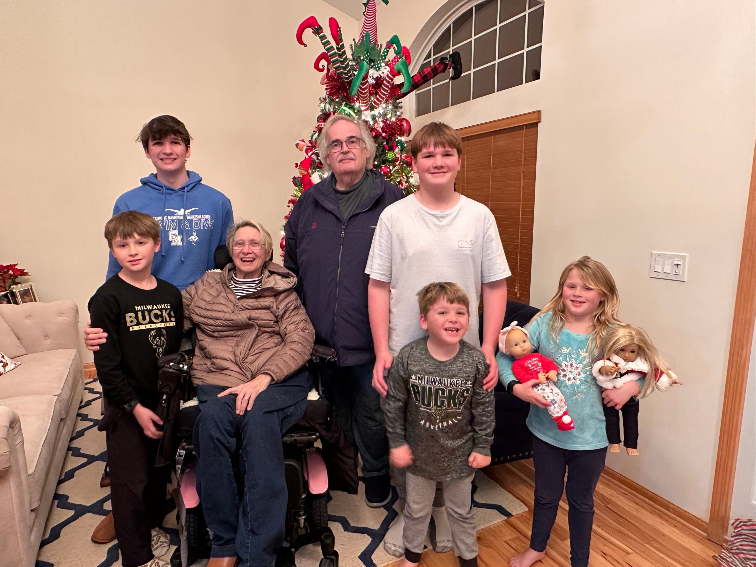 Group of children and adults posing for a Christmas photo indoors, with a decorated Christmas tree in the background.