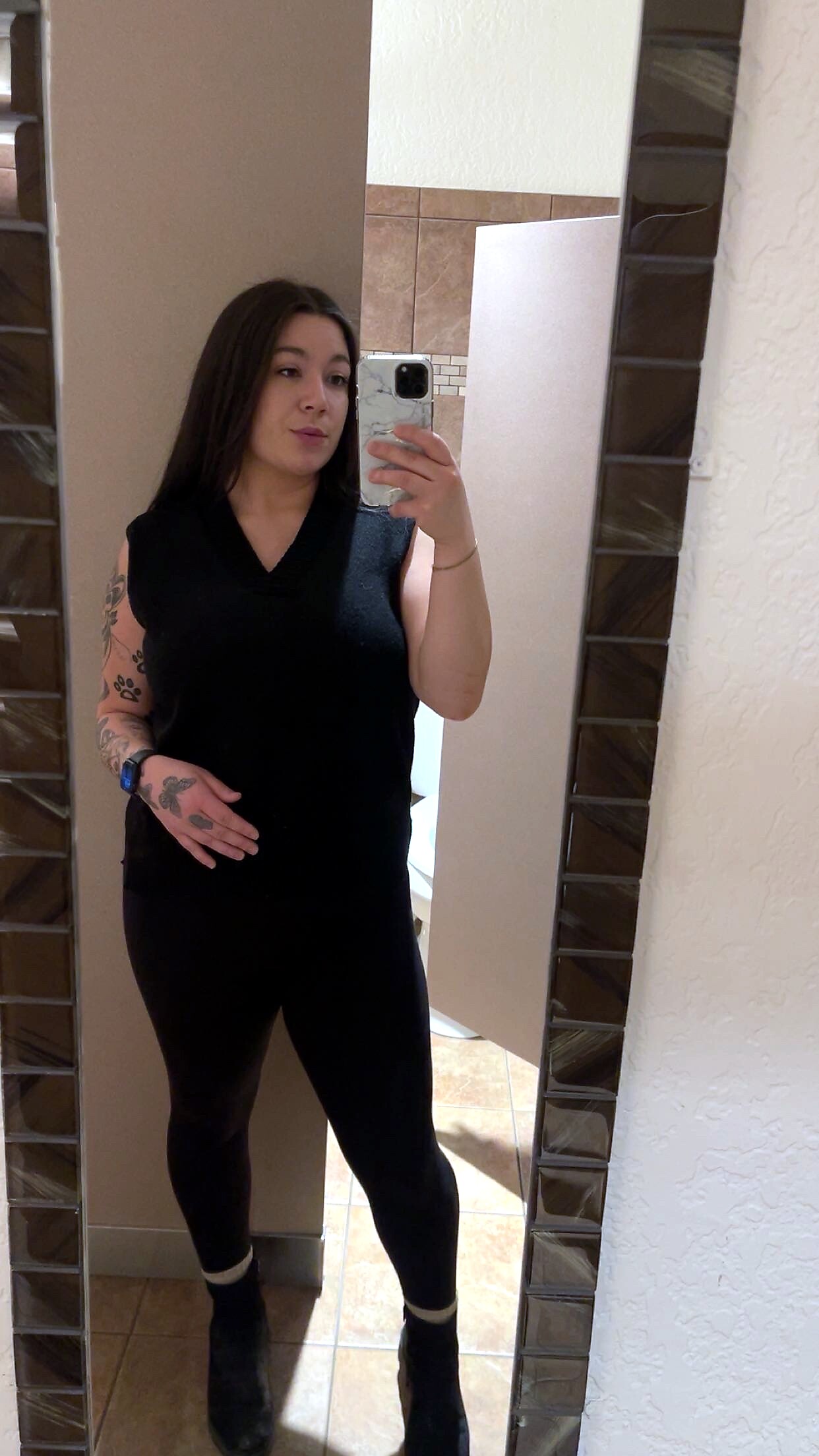 Woman with dark hair wearing a black sleeveless top and black leggings taking a mirror selfie in a tiled bathroom; tattoos on her left arm visible.