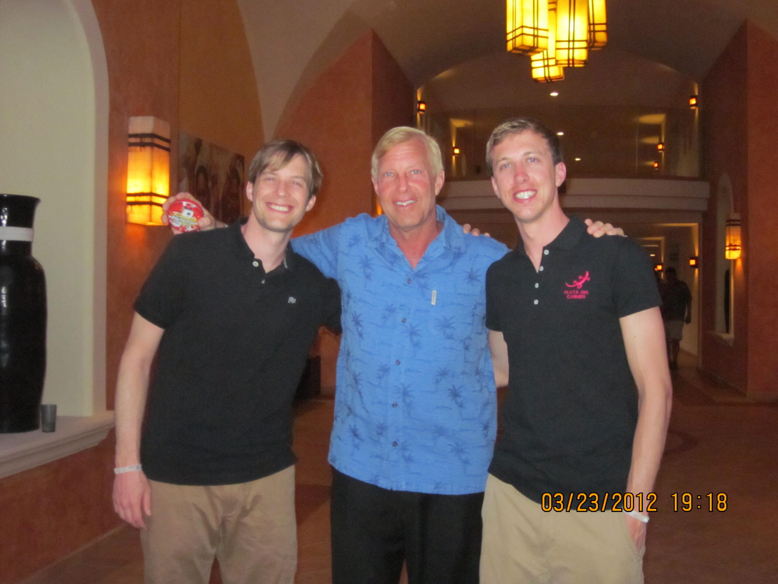 Three men posing together indoors; center man in a blue tropical shirt, flanked by two men in black polo shirts.