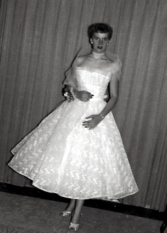 Woman in a vintage mid-20th-century ball gown, posing with hands on her skirt in front of curtains (black-and-white photo).