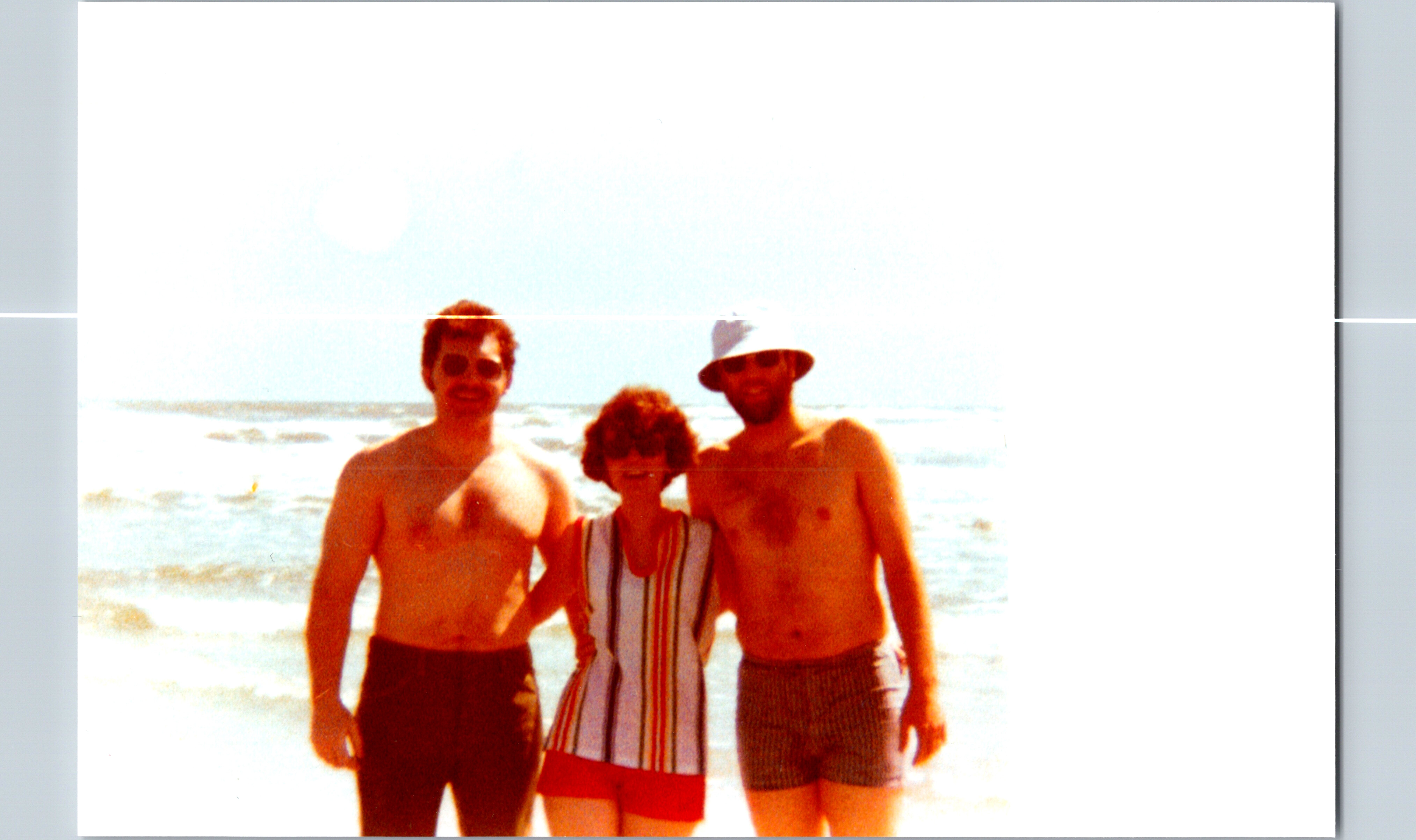 Three friends stand together on a sunny beach, smiling in swimsuits with the ocean in the background.