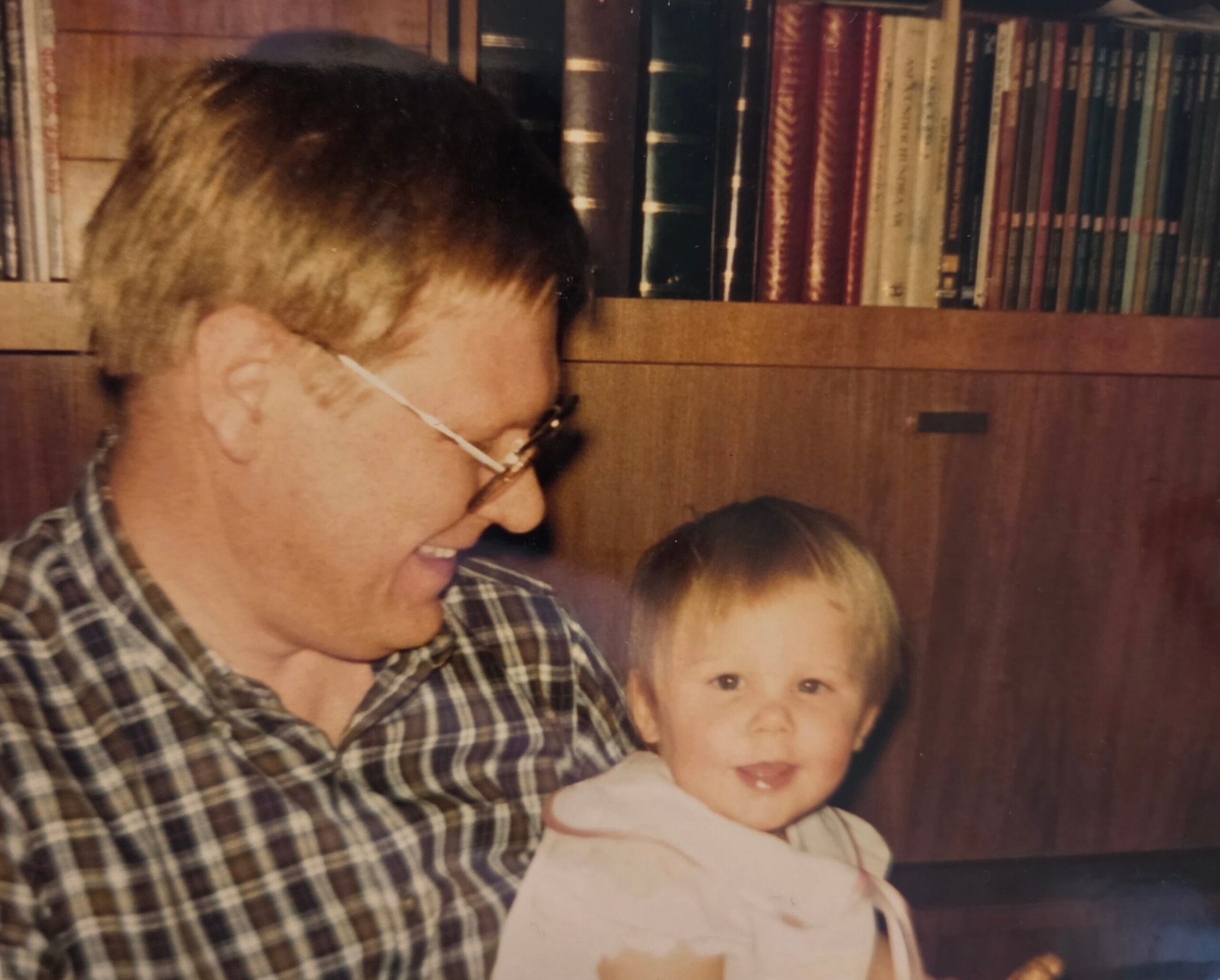 Adult with glasses and a plaid shirt holding a smiling baby, with a wooden bookshelf full of books in the background.