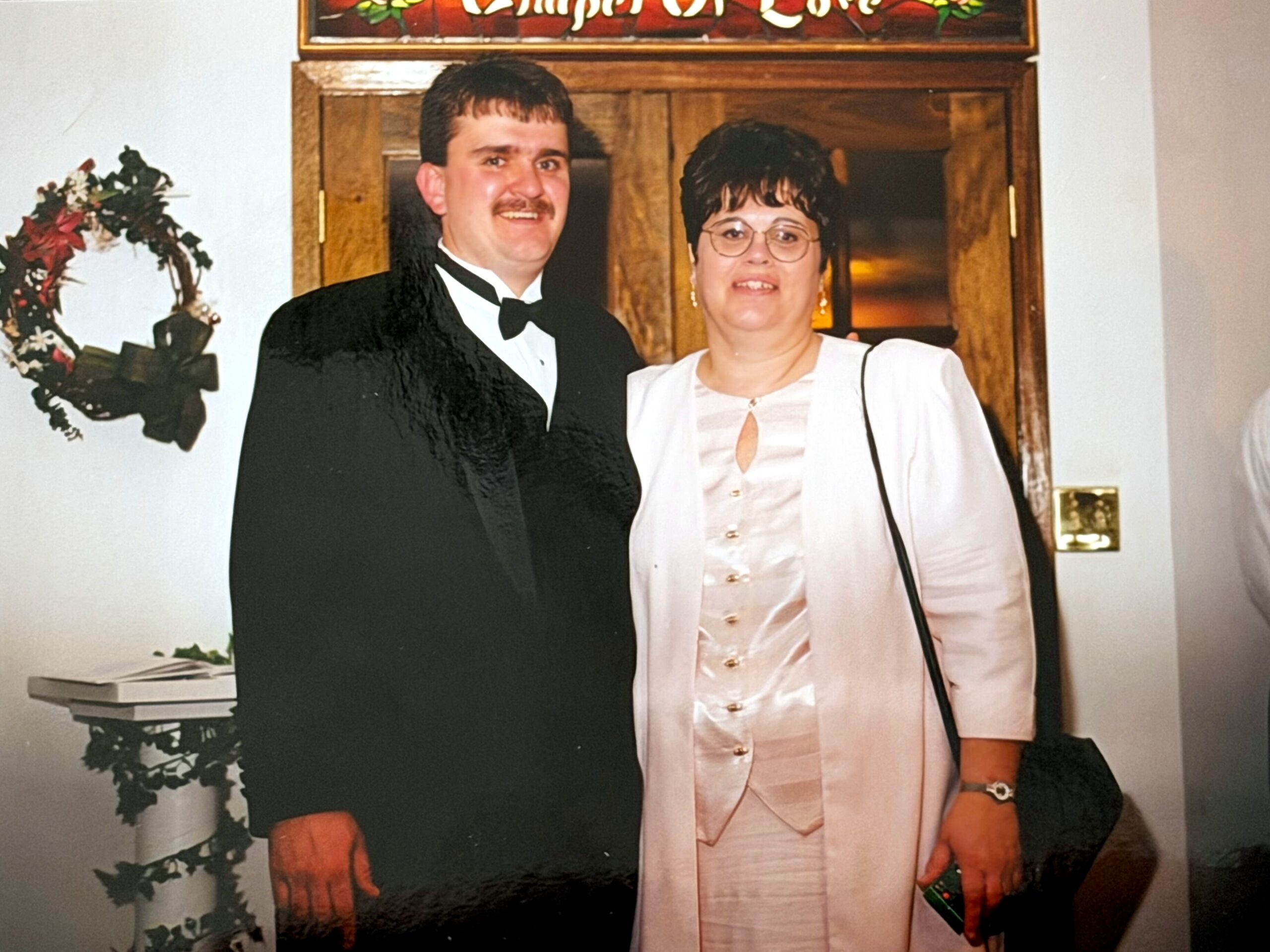 A couple posing for a photo at a formal event: man in a black tuxedo and bow tie, woman in a pale pink dress with glasses, standing by a wooden door with a wreath nearby.