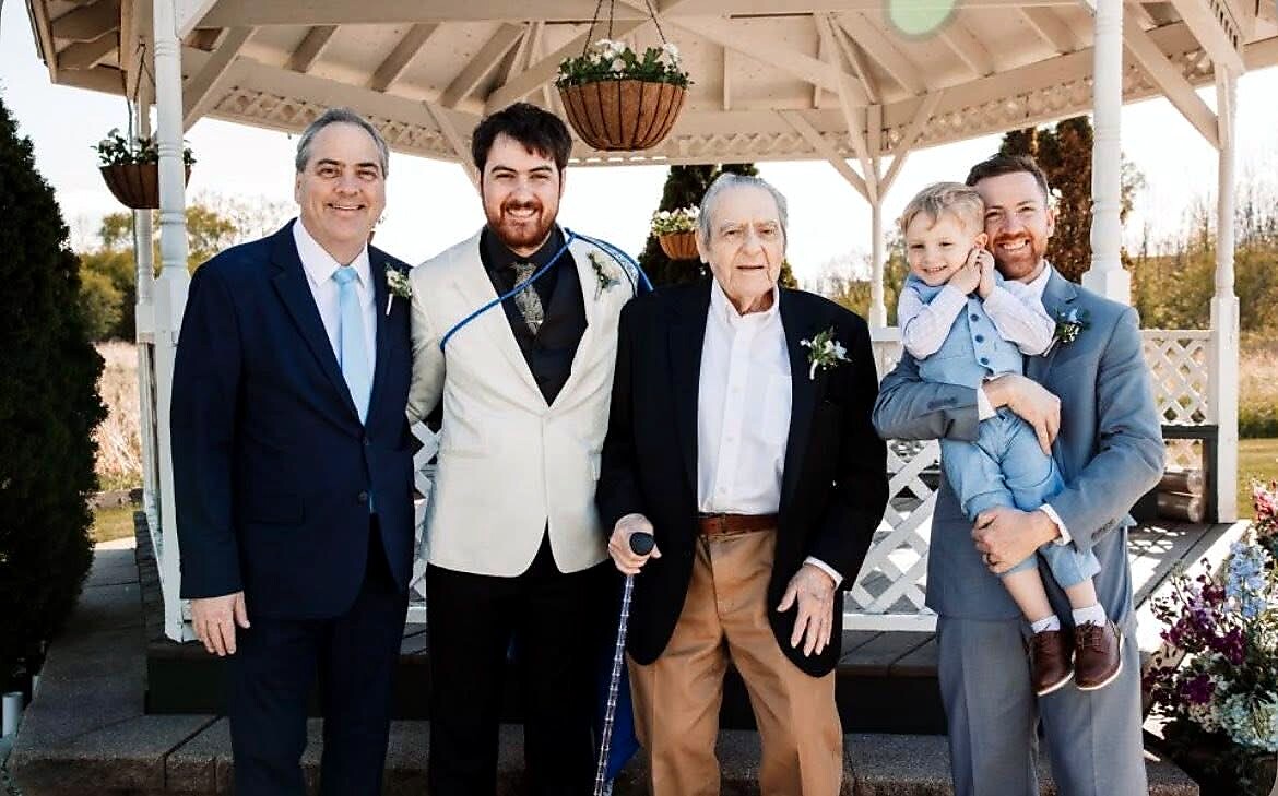 Group of five men posing together outdoors under a gazebo; elder man with a cane stands center, flanked by four men in suits, one holding a toddler.