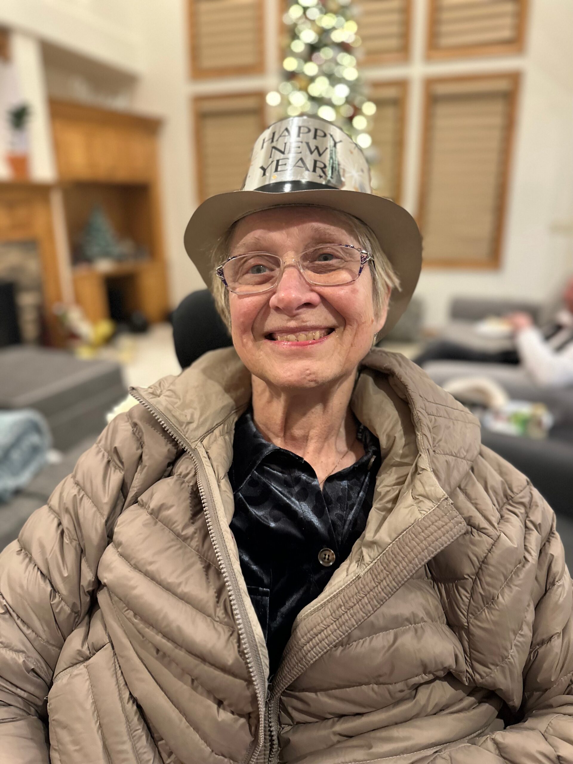 Smiling elderly woman in a beige quilted jacket and silver 'Happy New Year' hat indoors, with Christmas lights in the background.
