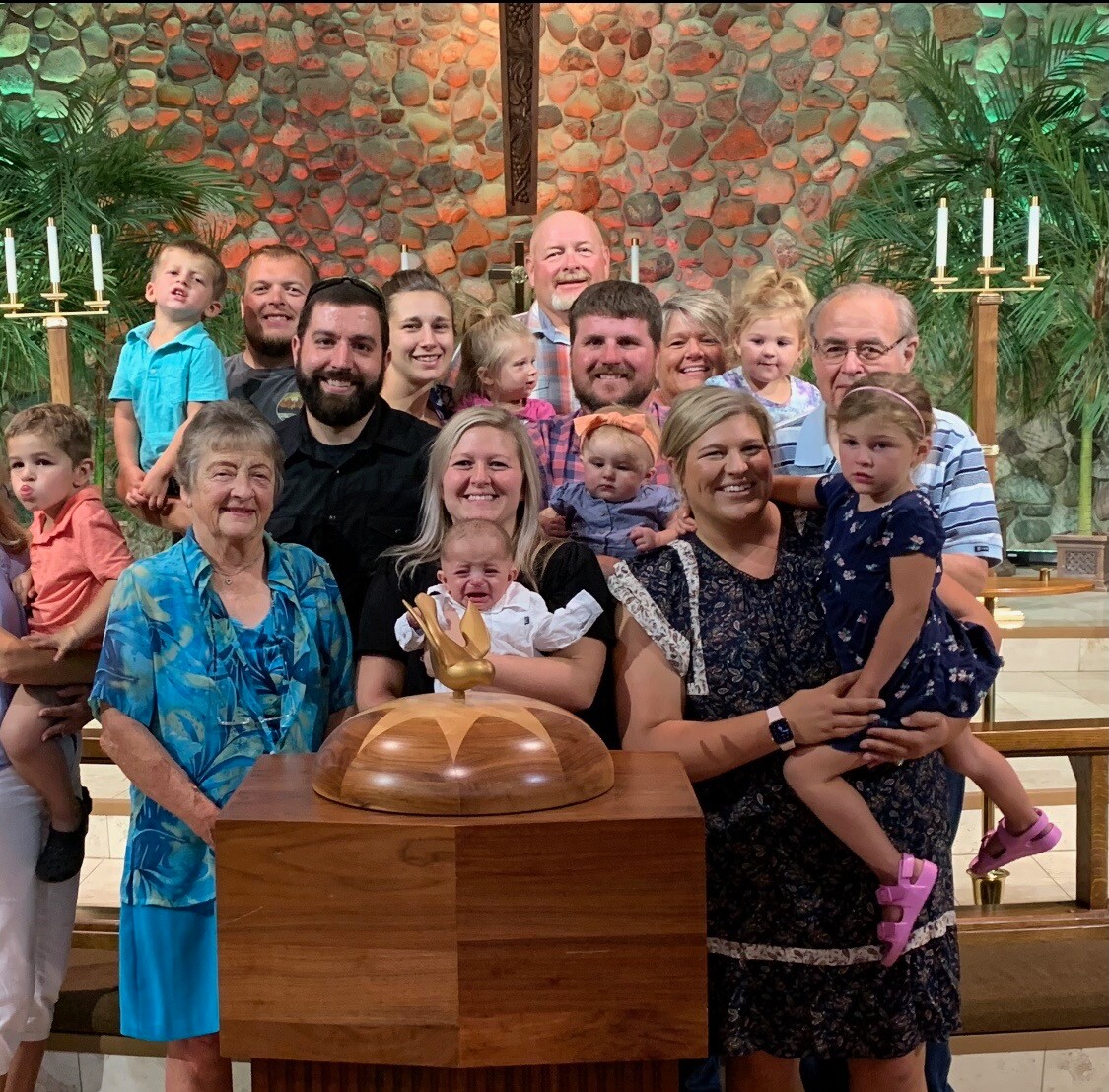 A multigenerational family posing for a group photo in a church, gathered around a wooden pulpit with a gold dove ornament, stone wall and candles in the background.