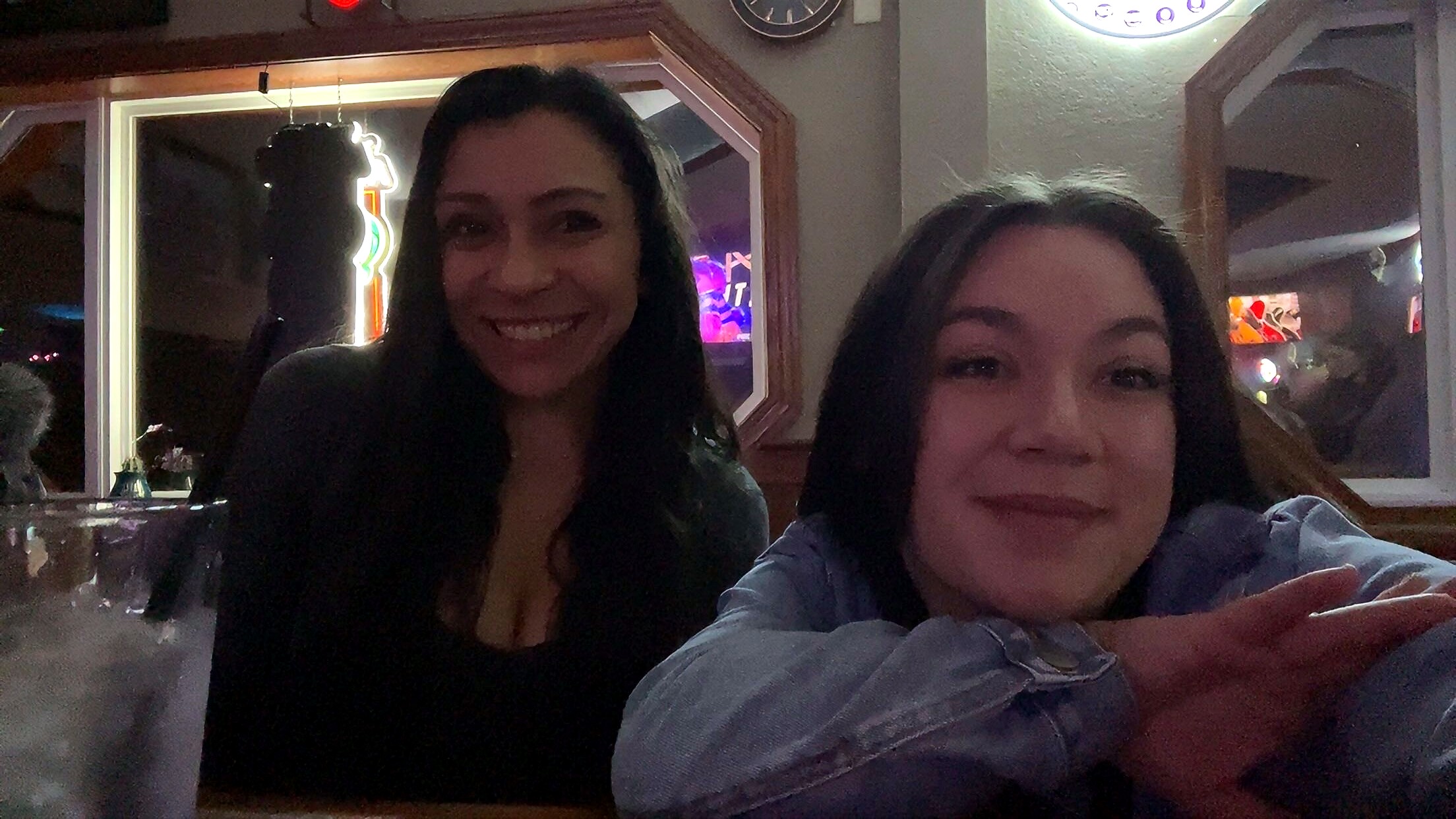 Two women smile for a selfie in a dimly lit bar, with neon lights and a window in the background.