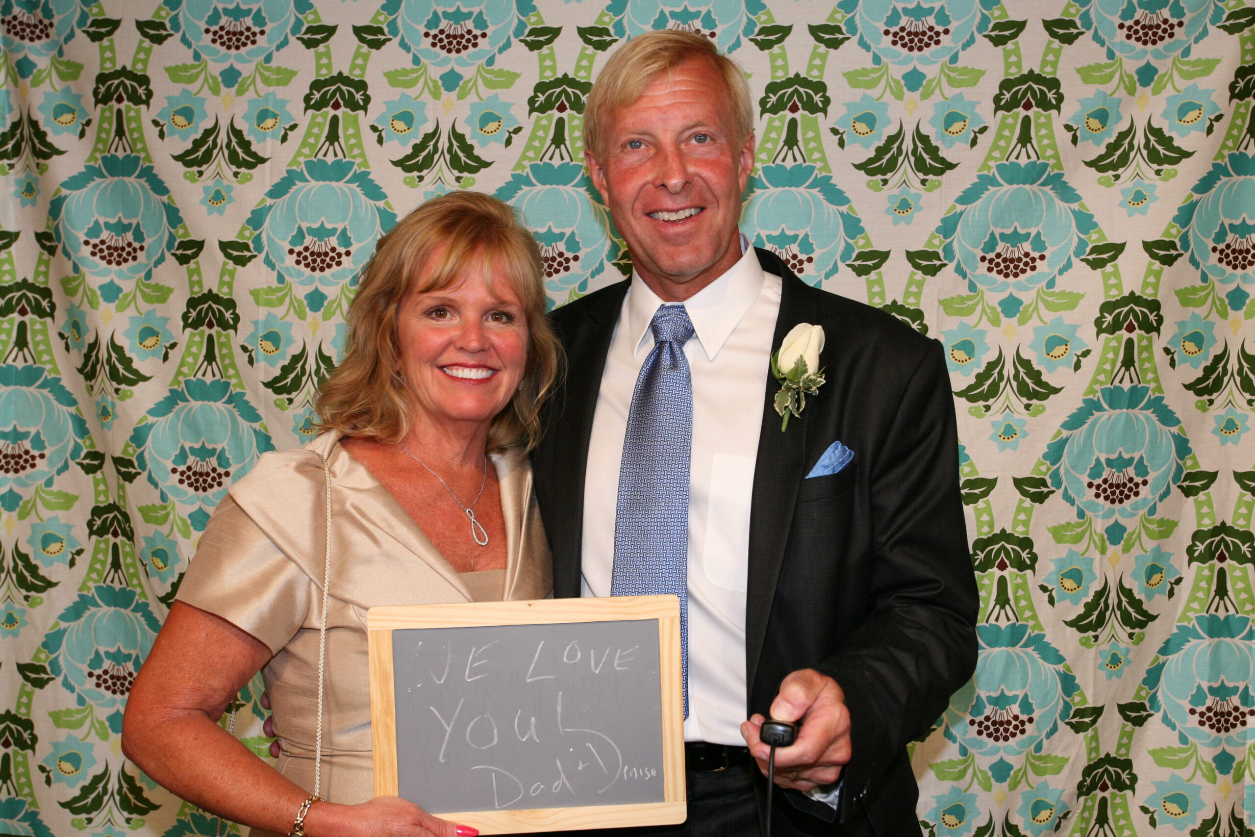 Smiling couple stands together in front of a bright patterned backdrop; the woman holds a chalkboard that reads 'WE LOVE YOU DAD'.